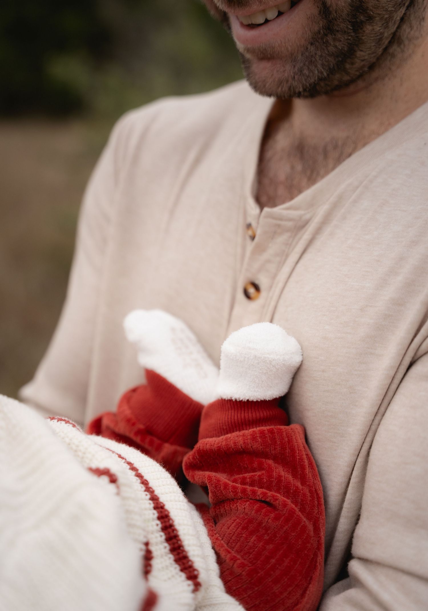 Father in beige henley shirt smiling while holding newborn baby in red and white outfit.