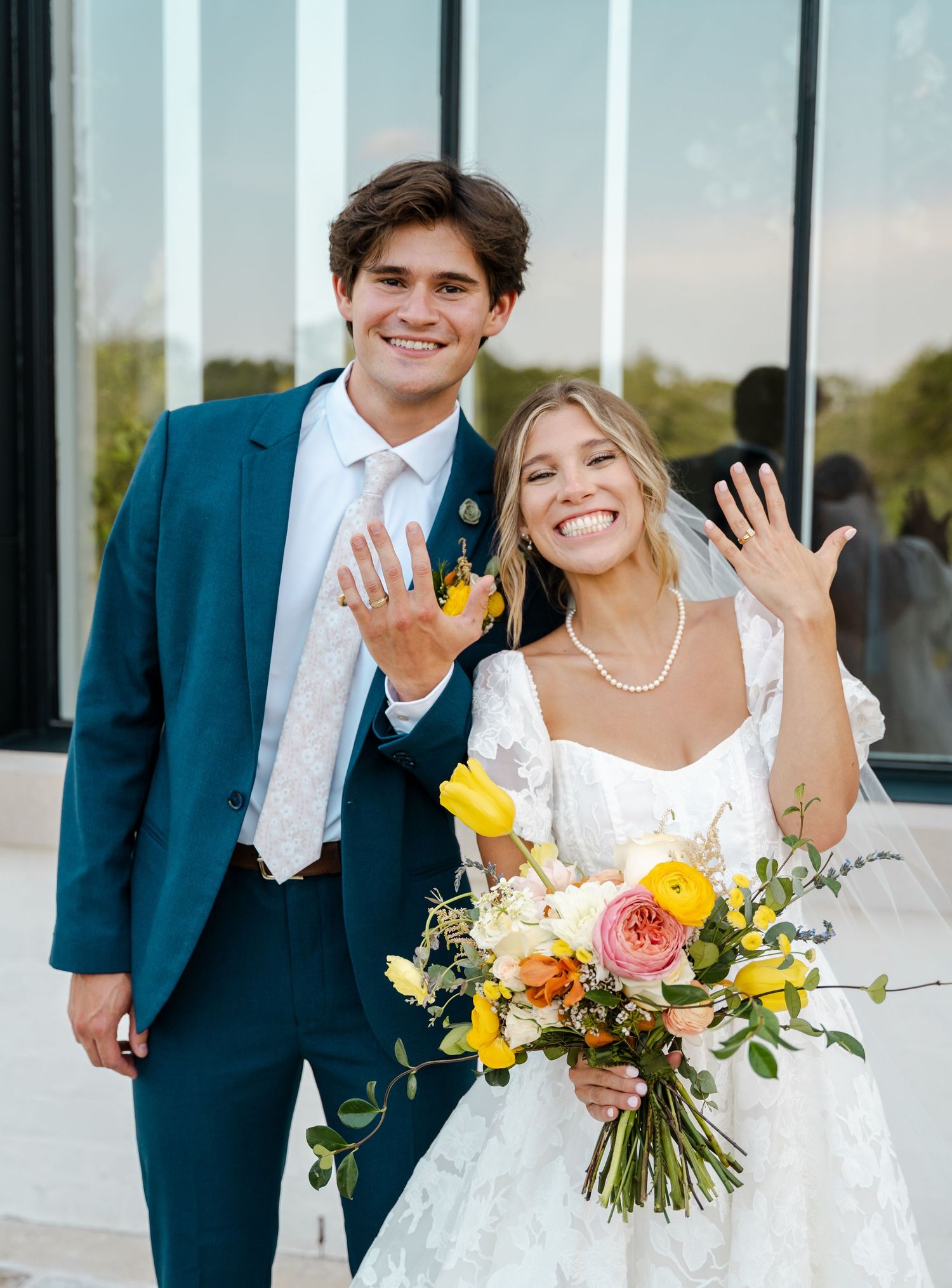 Smiling newlyweds showing off wedding rings, groom in teal suit, bride holding yellow bouquet.