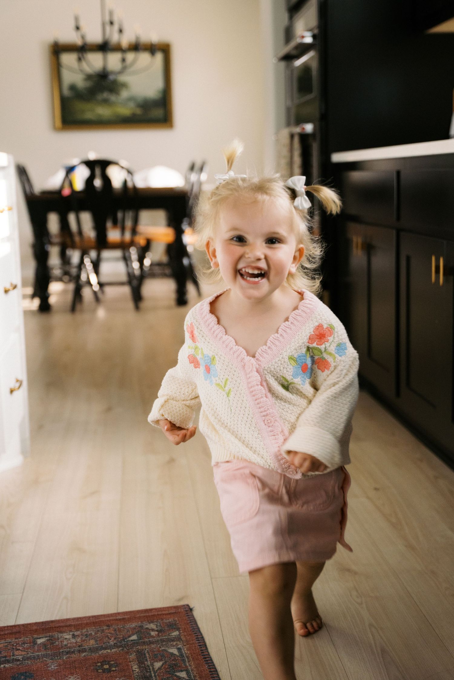 A happy toddler girl in a floral cardigan and pink skirt runs laughing through a kitchen.