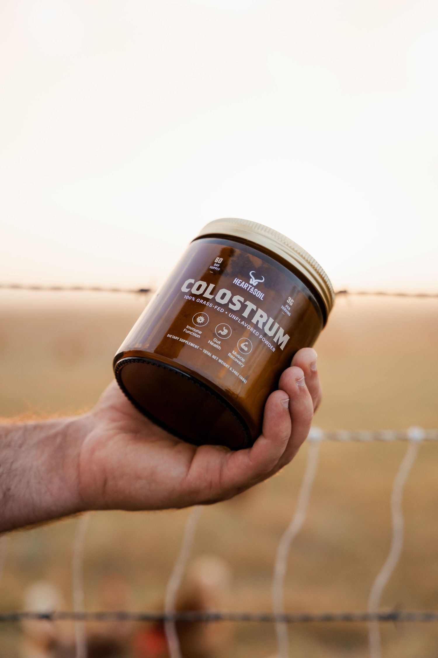 Hand holding a jar of Colostrum supplement near a barbed wire fence outdoors.