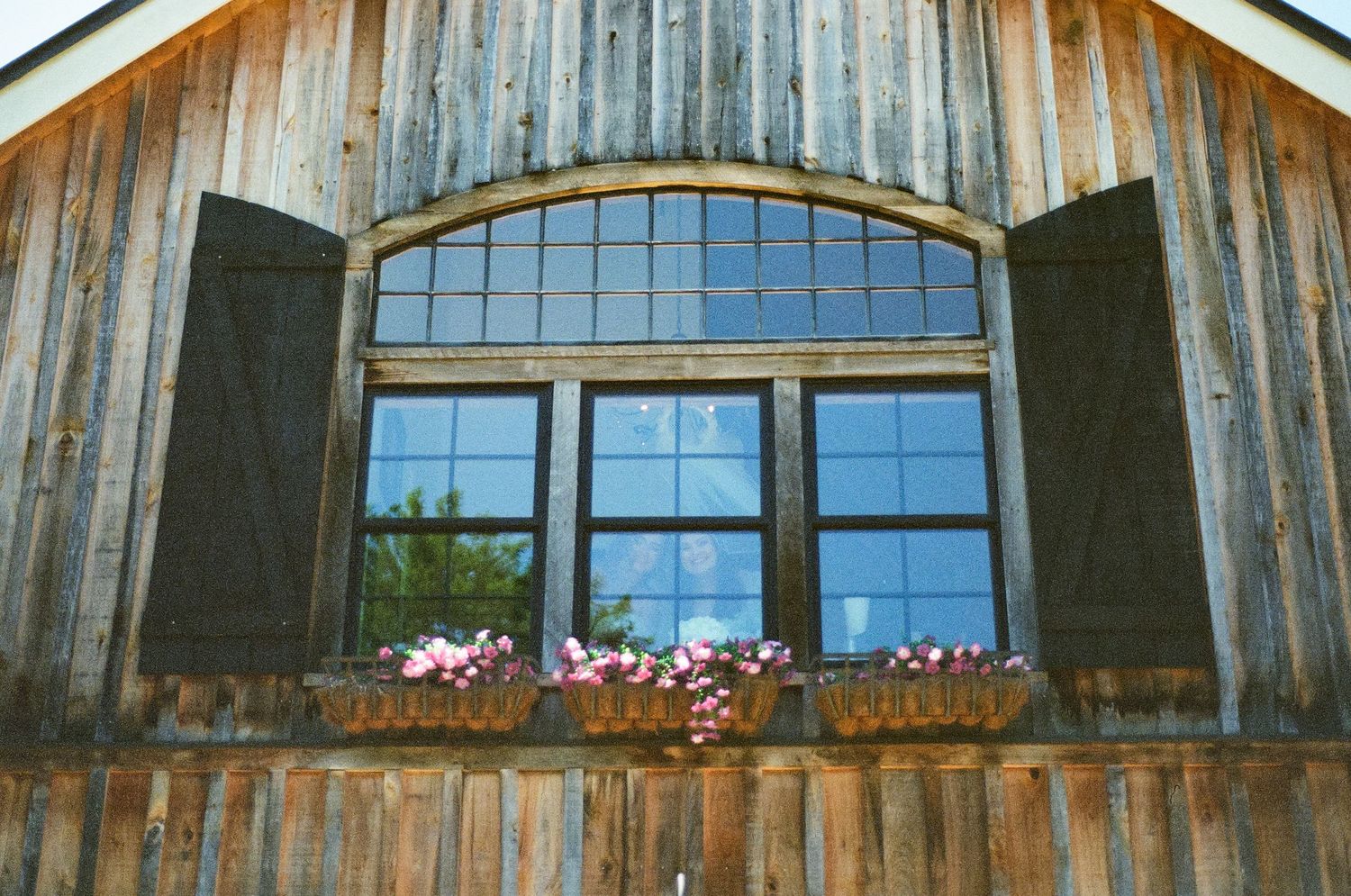Rustic wooden barn window with arched frame, dark shutters, and colorful flower boxes below.