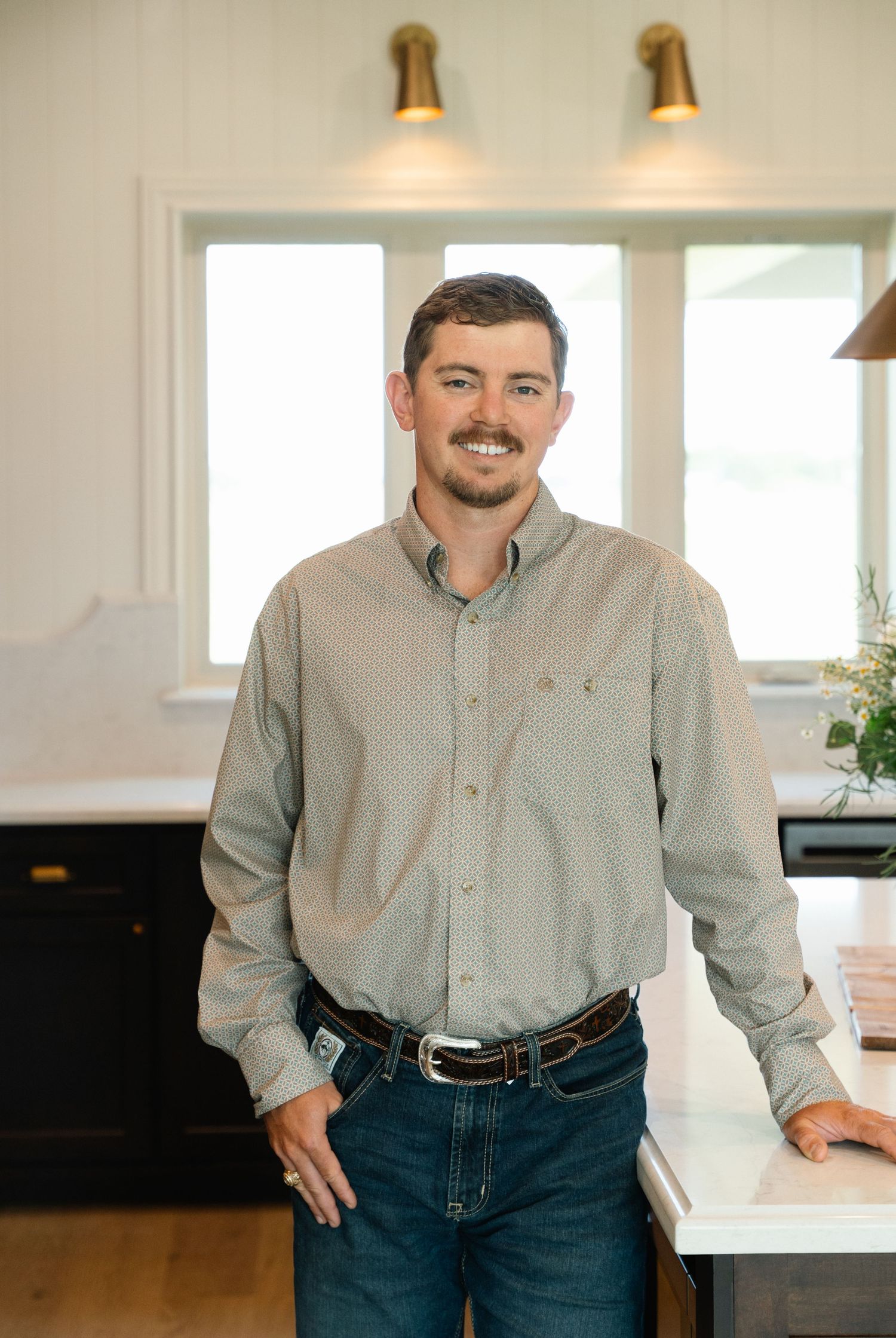 Smiling man in gray button-down shirt leaning on white marble kitchen counter in a bright modern home.