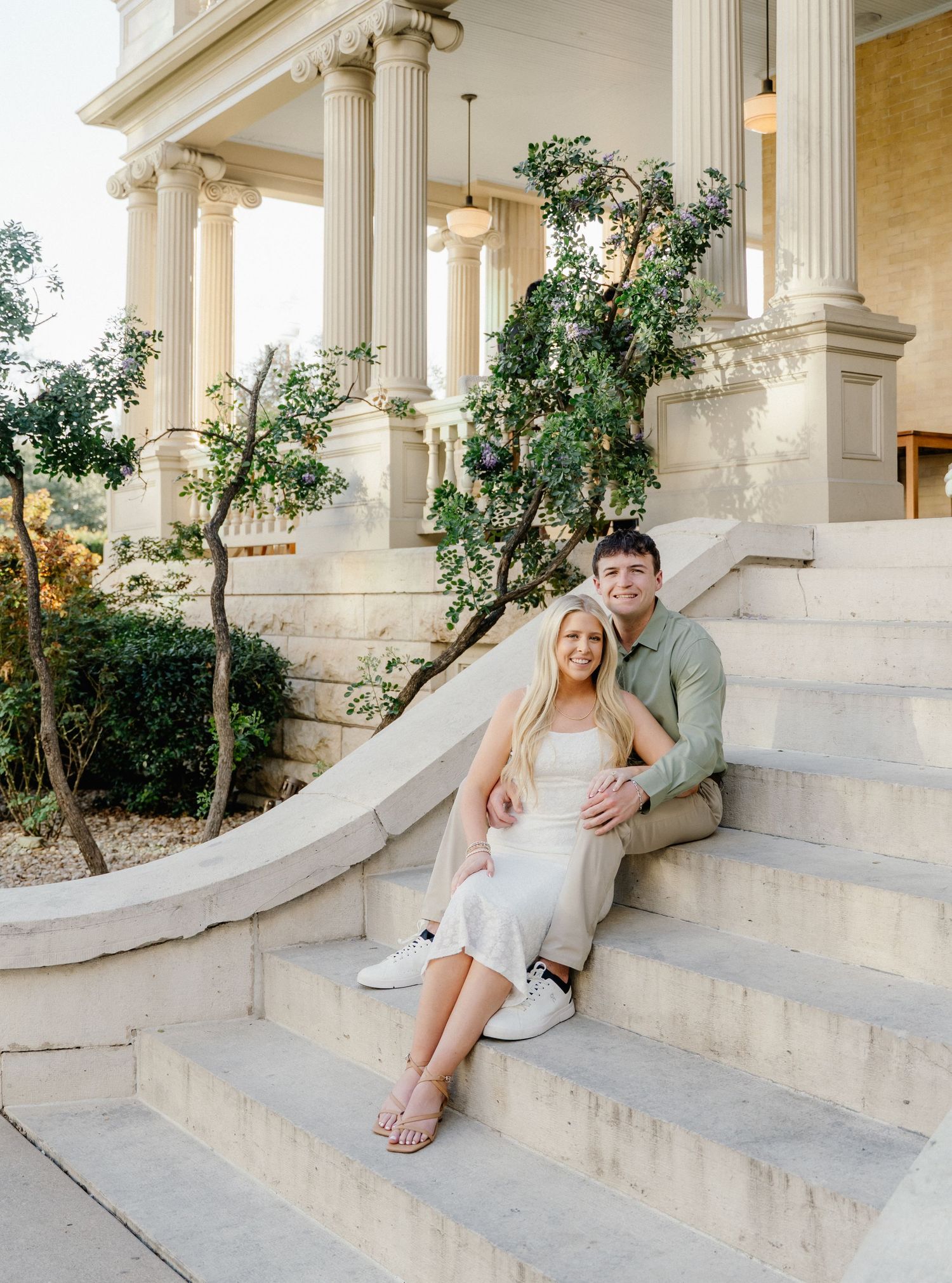 Couple sitting on stone steps of elegant columned building, woman in white dress, man in green shirt, engagement photo.
