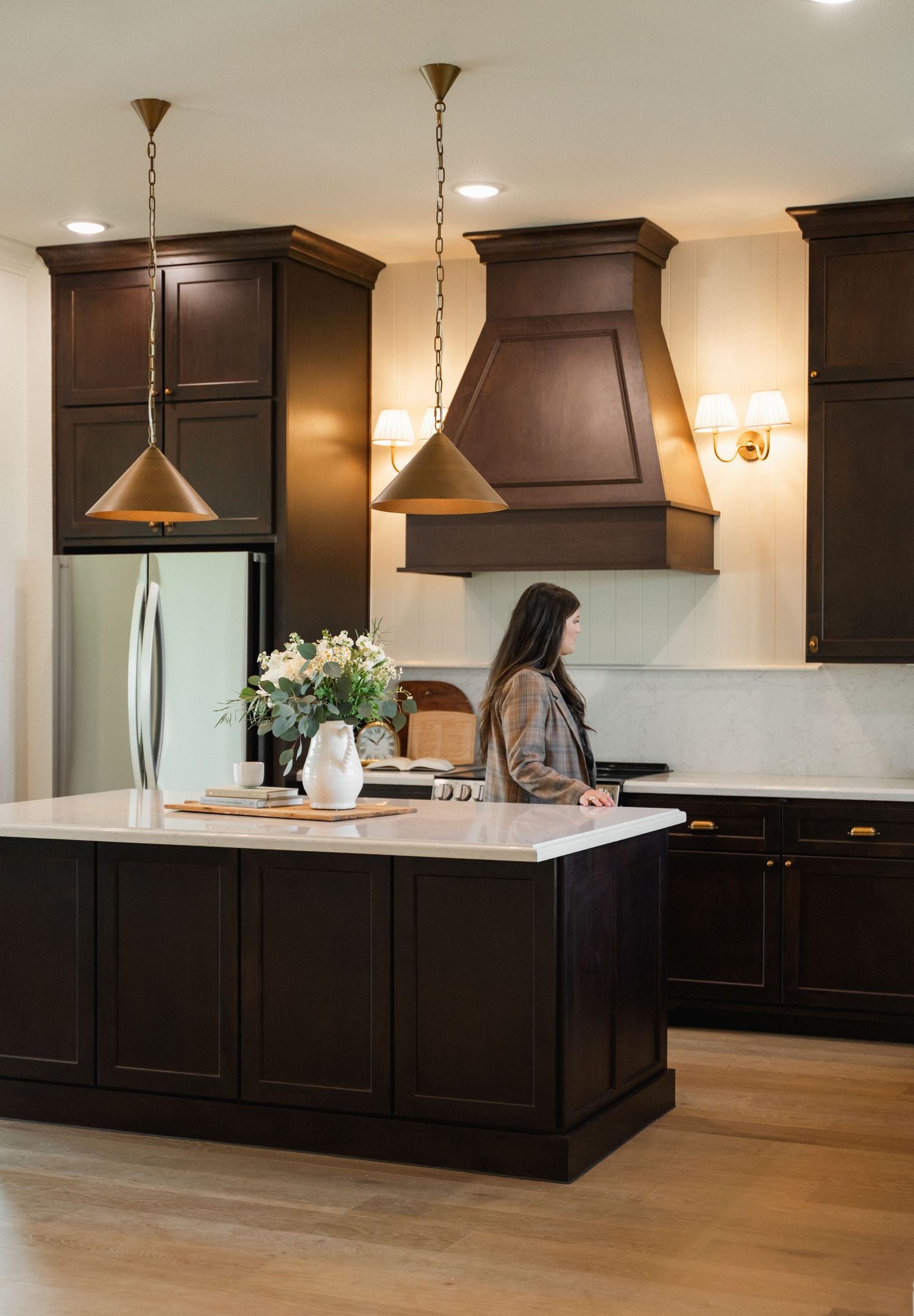 Elegant dark kitchen with white marble island, copper range hood, brass pendant lights, and woman standing nearby.