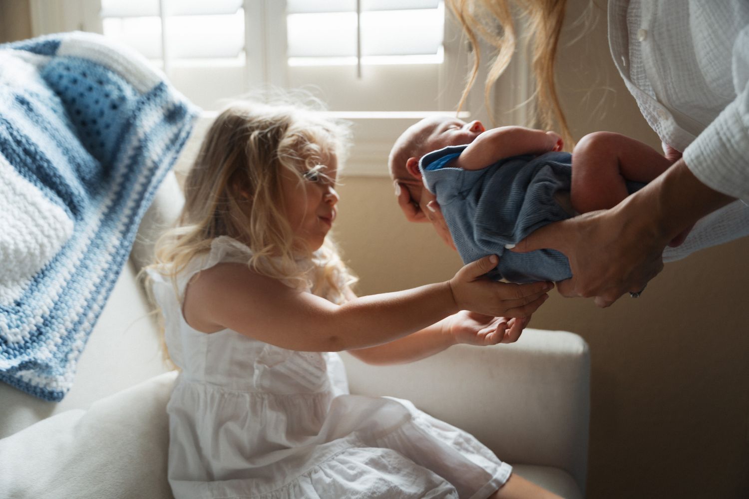 Young girl in white dress reaches out to touch newborn baby held by parent.