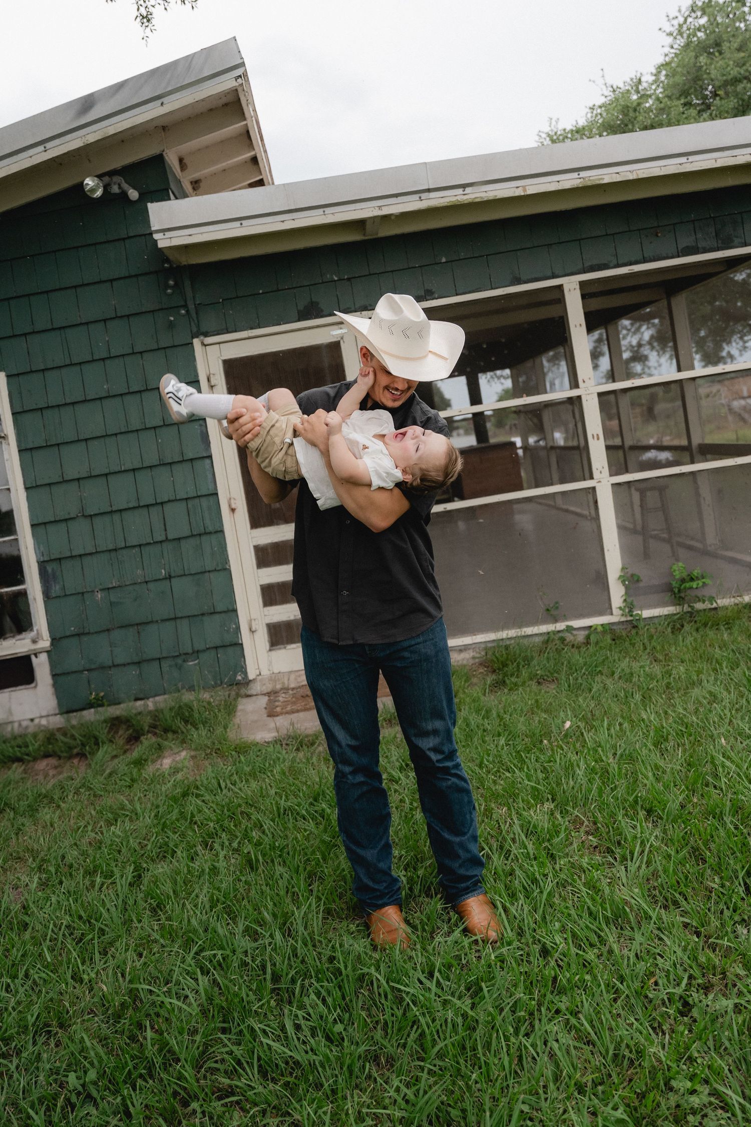 A father in a cowboy hat joyfully lifts his laughing baby outside a green house.