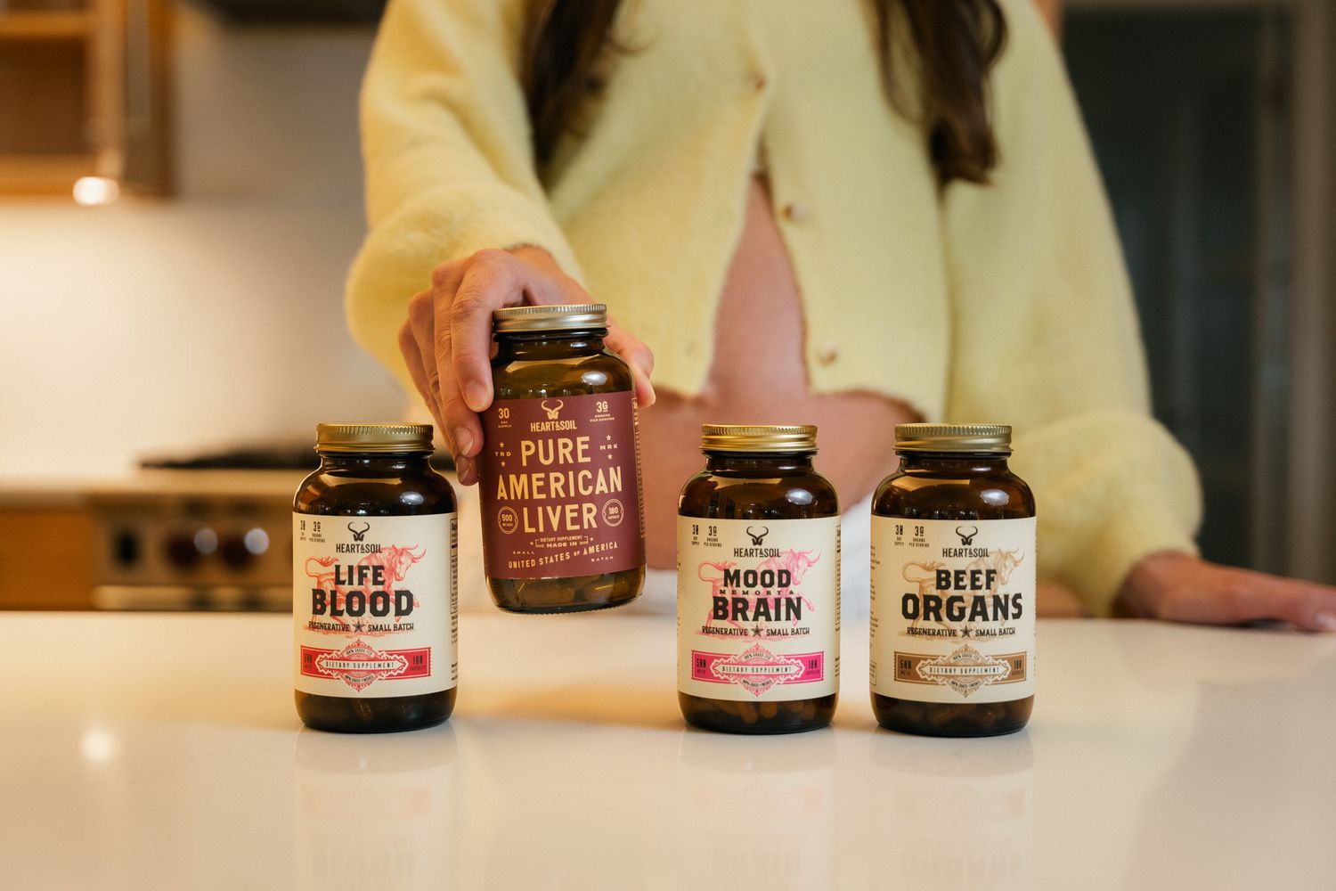 Woman holding Pure American Liver supplement with Life Blood, Mood Brain, and Beef Organs bottles on white counter.