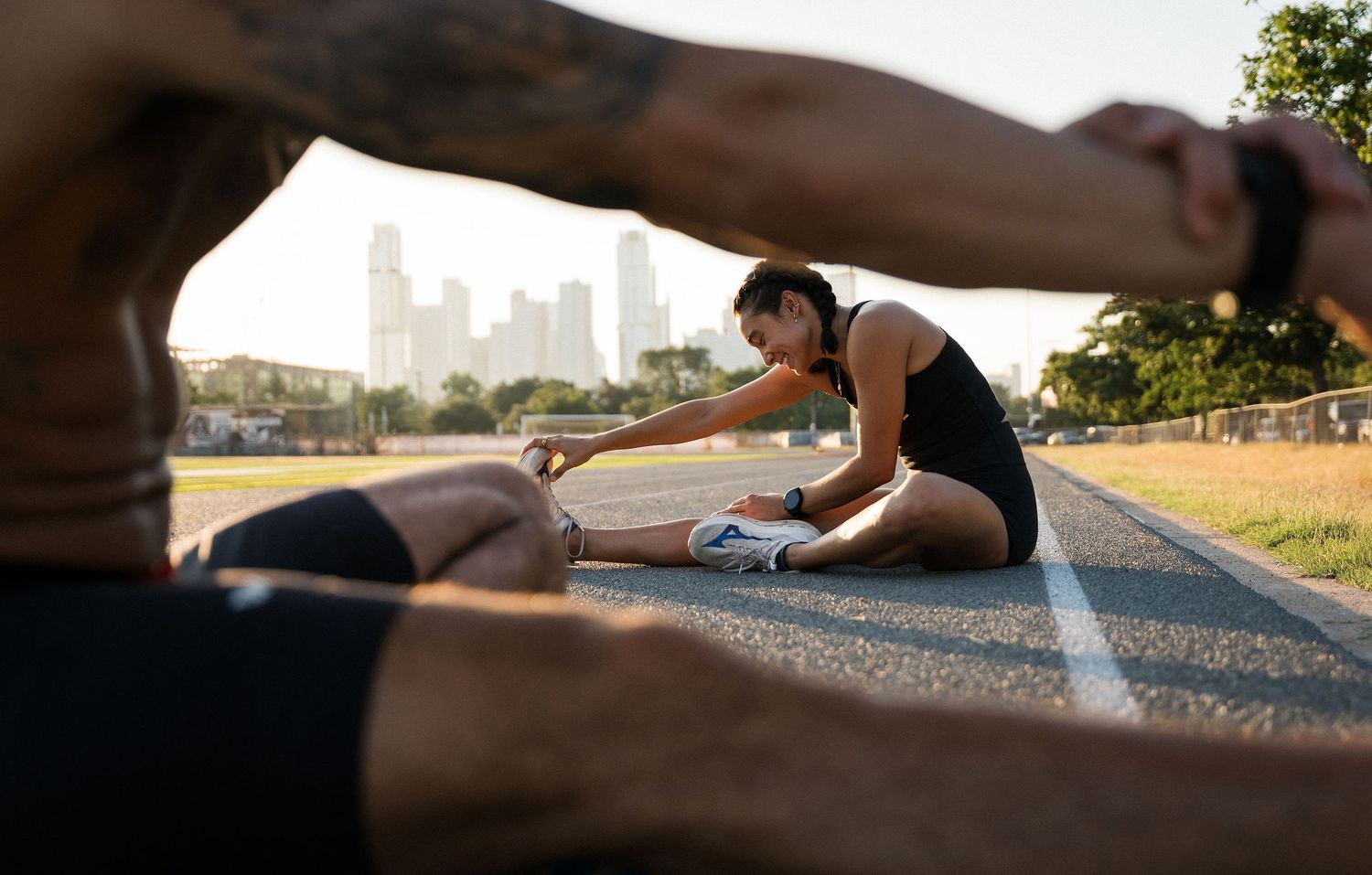 Two athletes stretching on an outdoor track with a city skyline in the background on a sunny day.