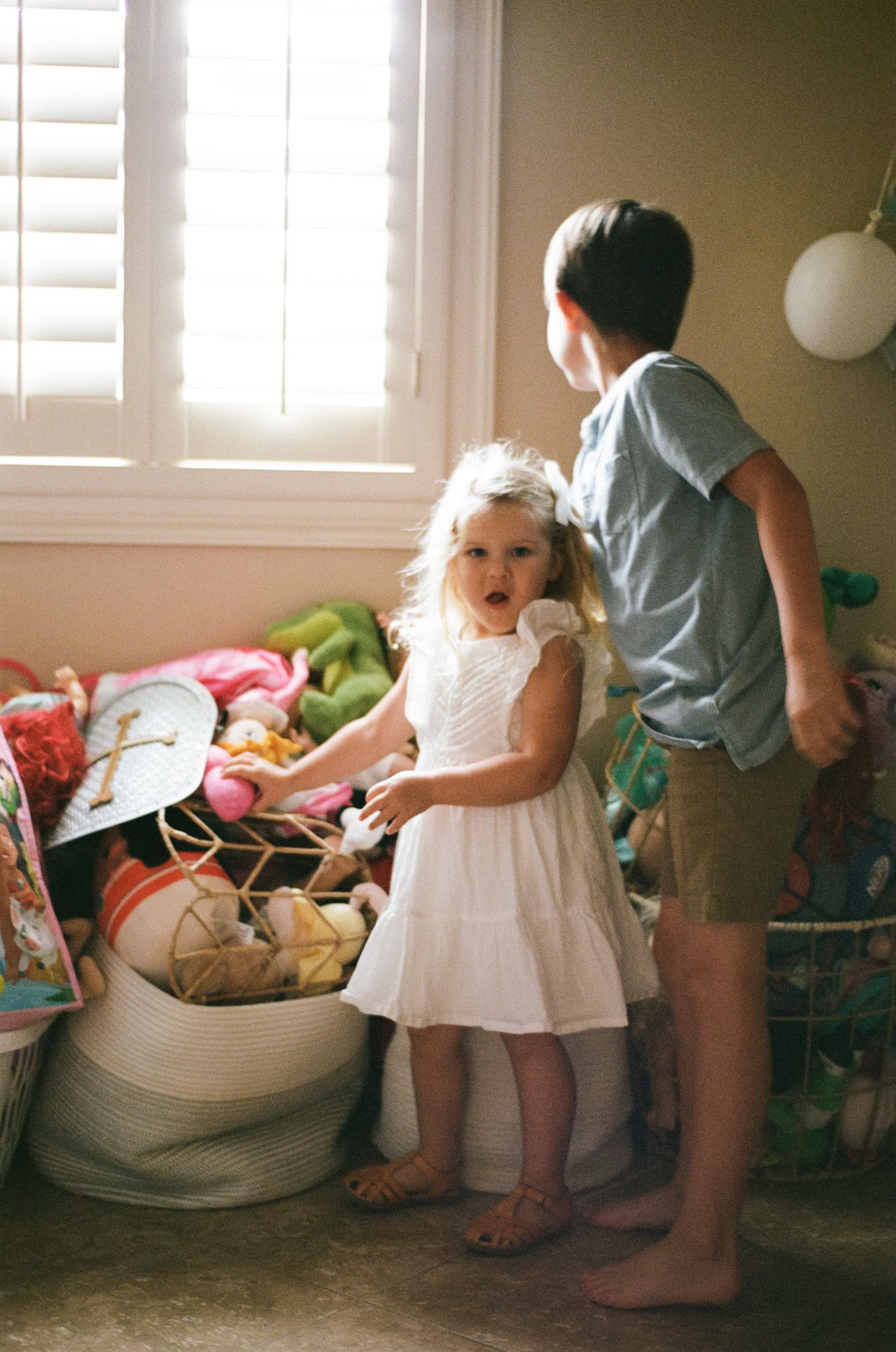 Two young siblings standing by window looking at toys in bright playroom.