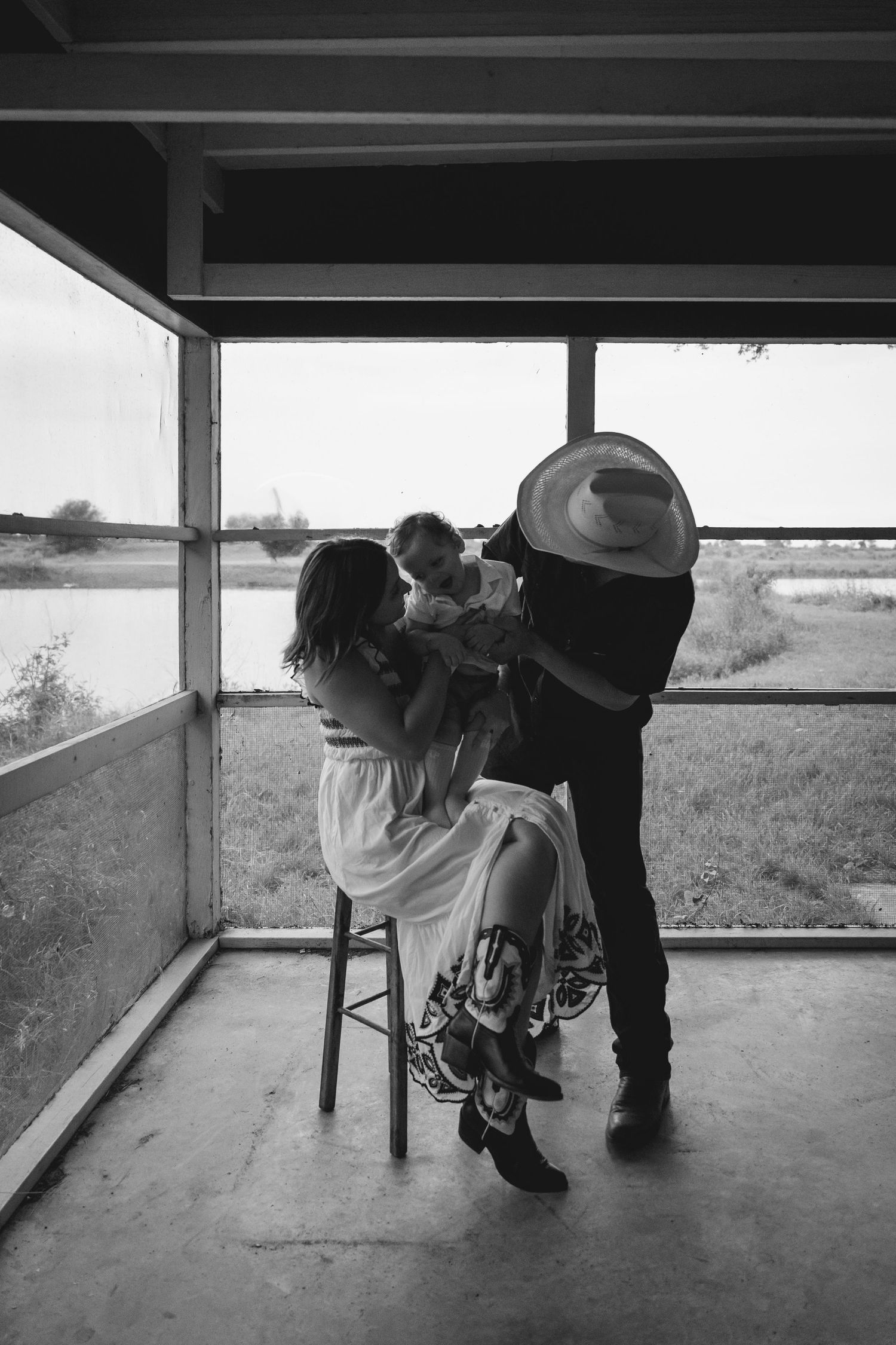 Black and white photo of a family with a cowboy hat-wearing father and mother holding their baby on a stool by a window.