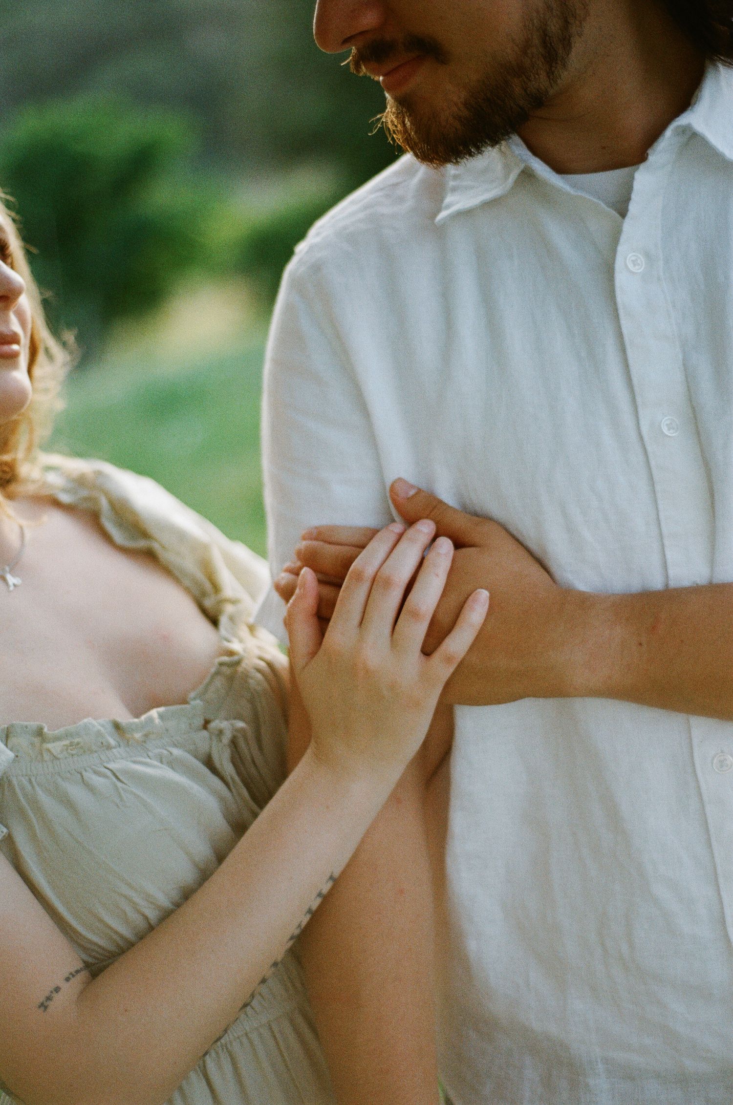 Couple holding hands tenderly in a sunlit garden, woman in sage dress and man in white linen shirt.