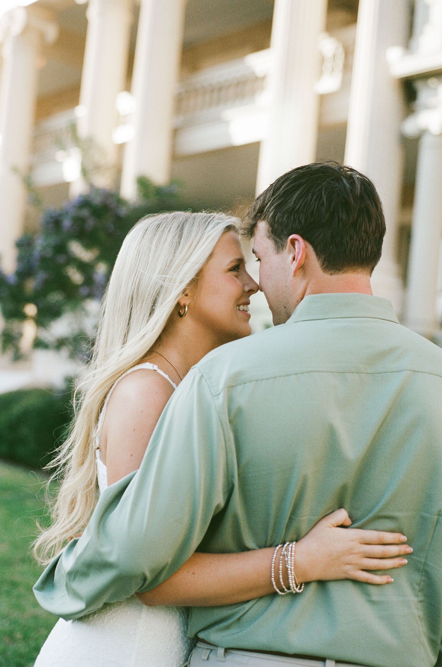 Smiling blonde woman embraces man in sage green shirt outside columned building in golden light.