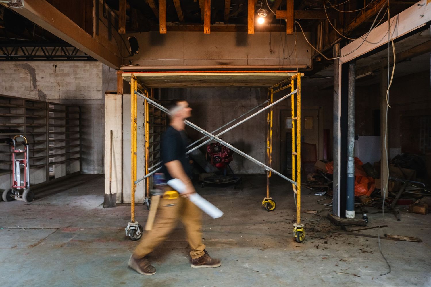 Construction worker carrying materials through an unfinished building interior with scaffolding and exposed wiring.