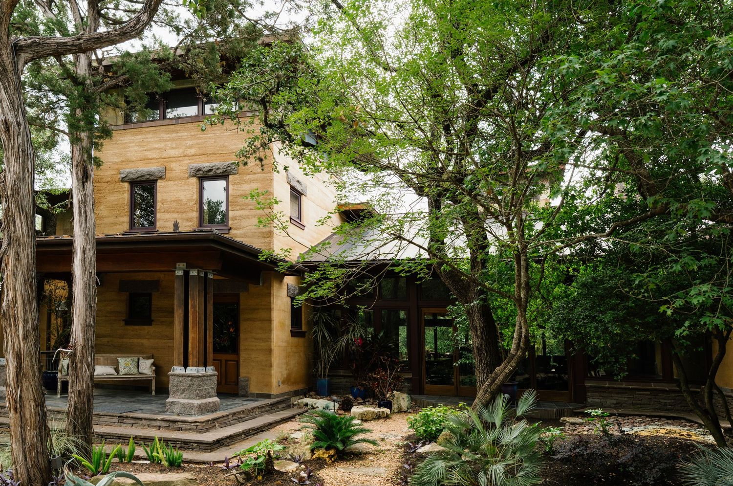 Unique rammed earth home nestled among mature trees with covered porch and natural desert landscaping.