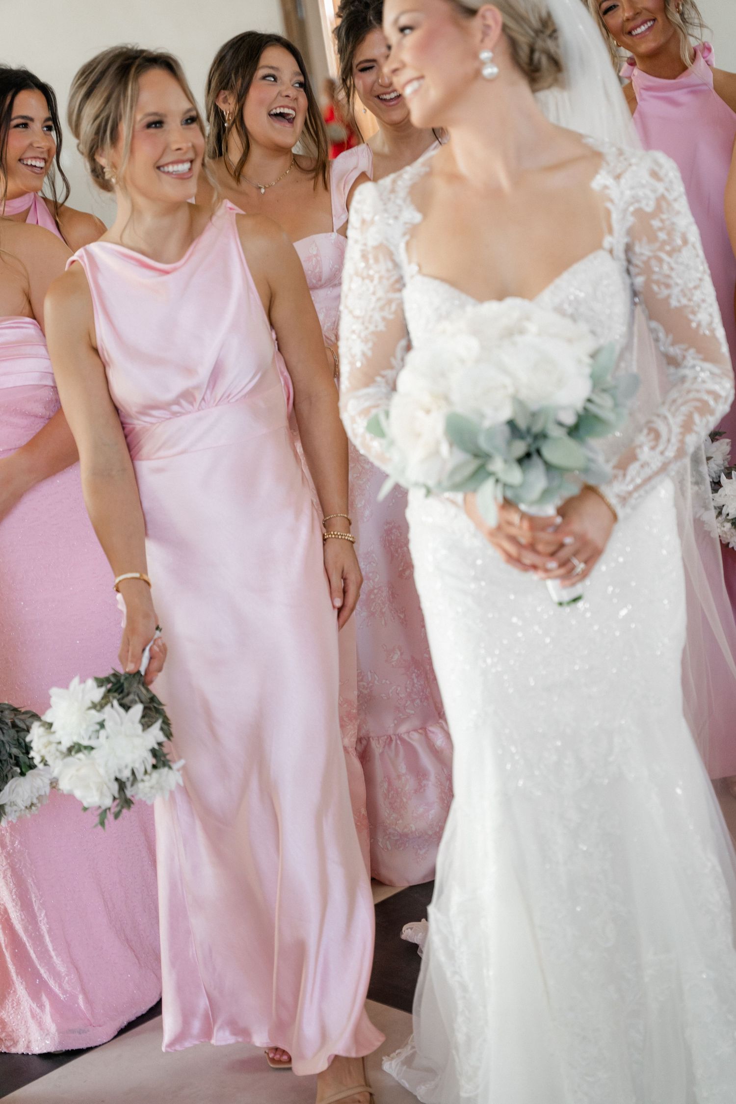 Bride in lace long-sleeve gown holding white bouquet laughing with bridesmaids in pink satin dresses.