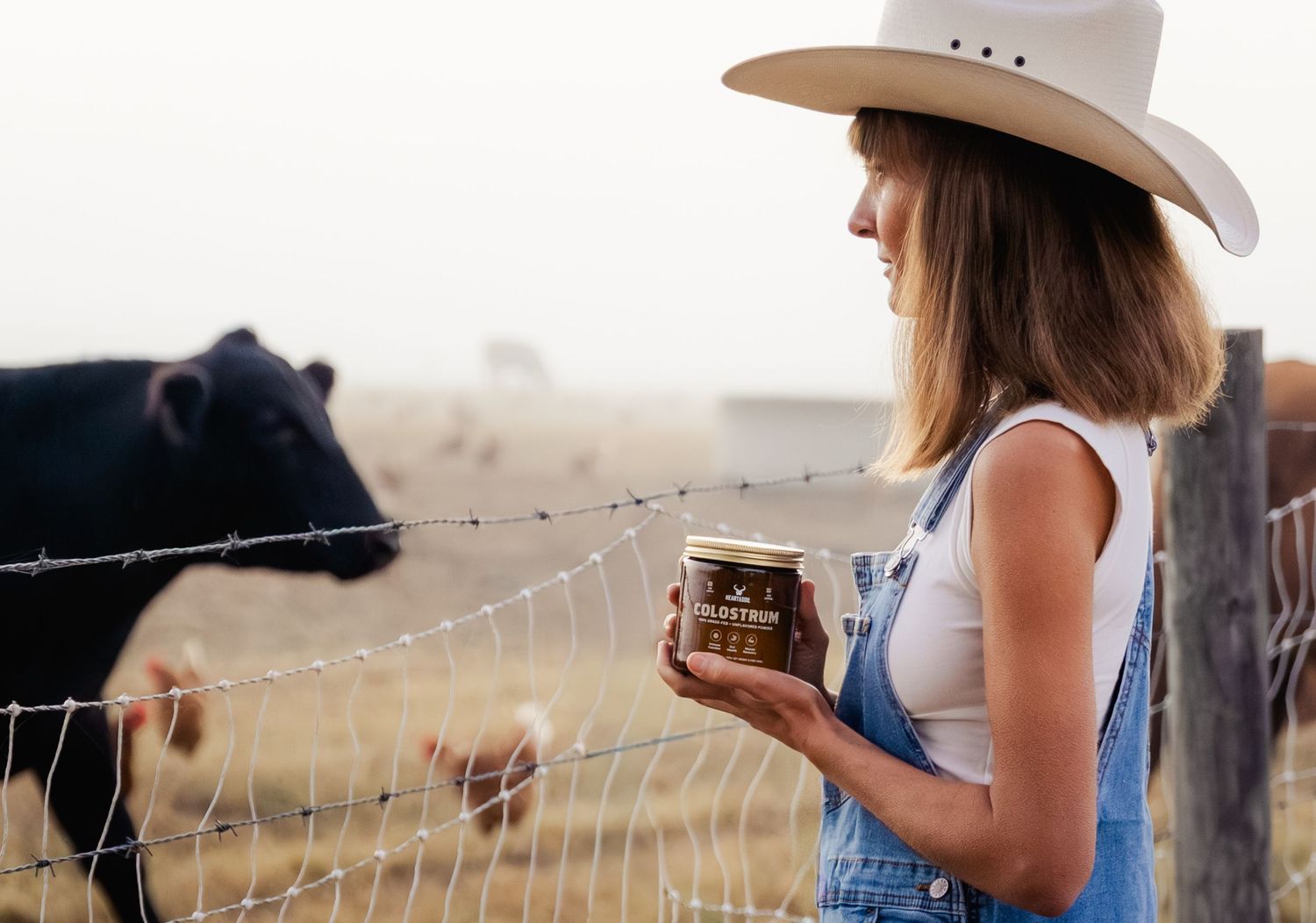 Woman in cowboy hat and overalls holding a jar near a black cow on a ranch.