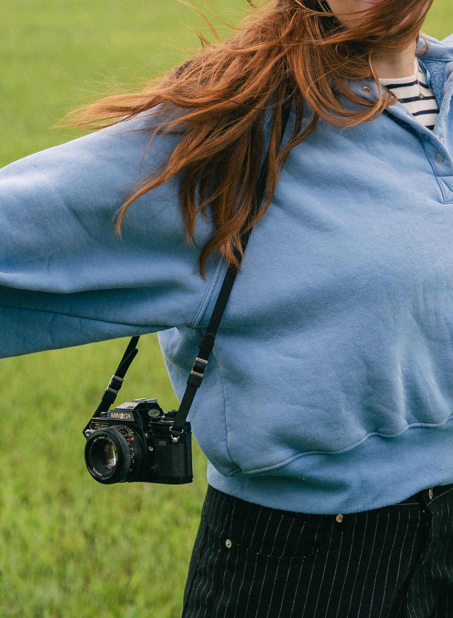 Red-haired woman in blue hoodie carrying a black film camera on a strap in a green field.