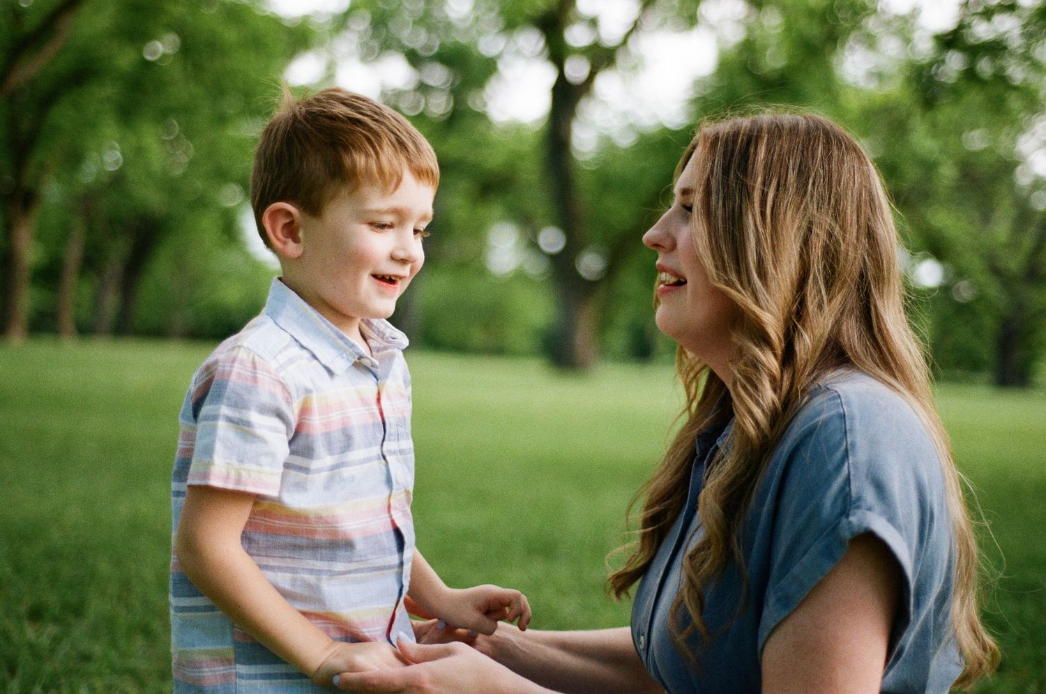 A smiling mother interacts joyfully with her red-haired toddler son outdoors.