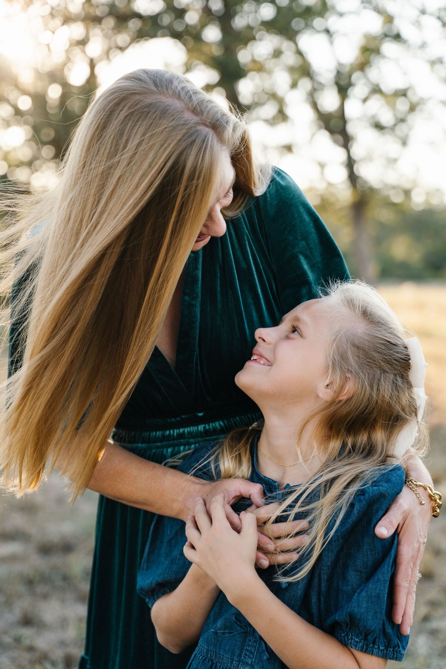 A mother in a green dress lovingly embraces her smiling blonde daughter outdoors.