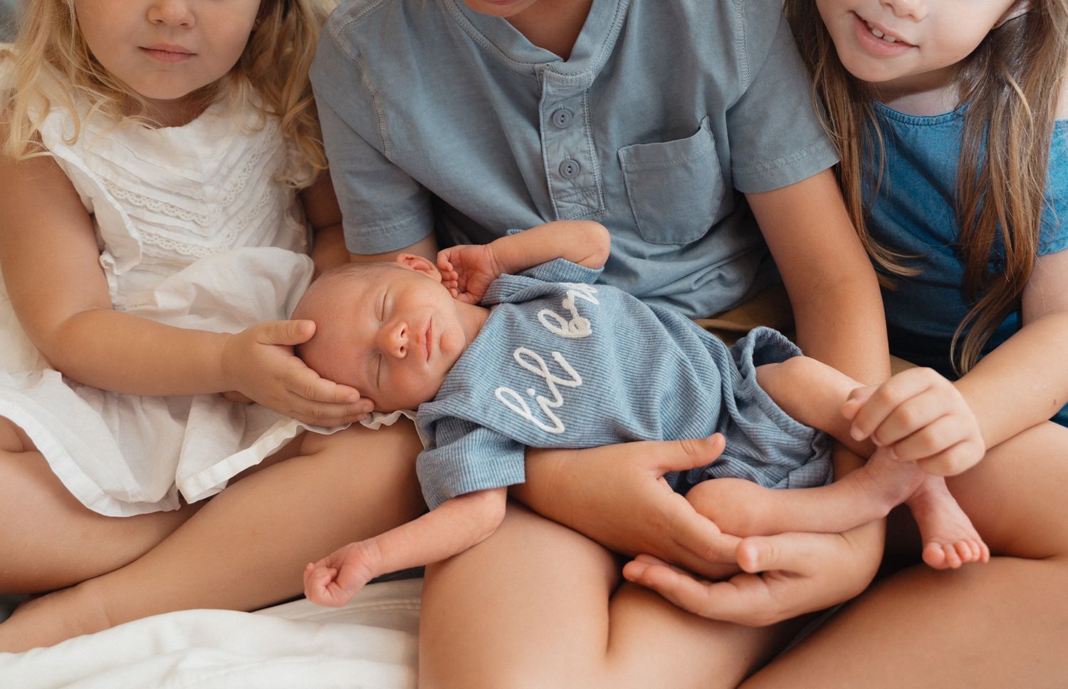 Three children gently hold a sleeping newborn baby dressed in a blue outfit.