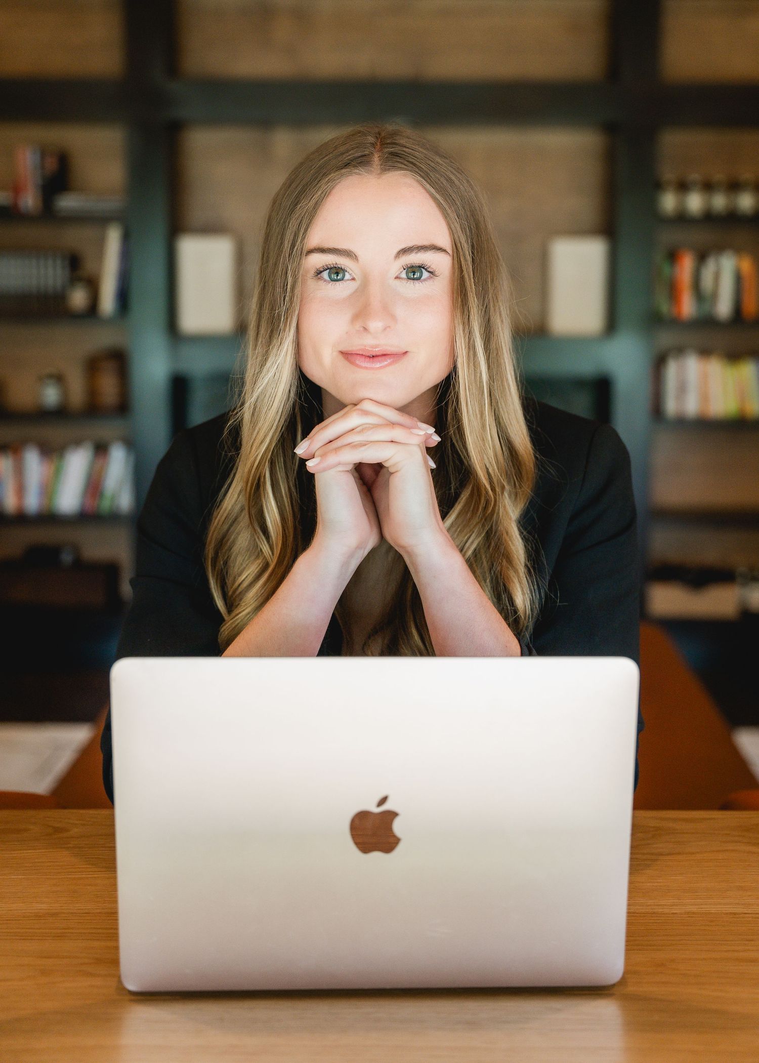 Young professional woman with blonde hair smiling at desk with MacBook laptop in modern office library setting.
