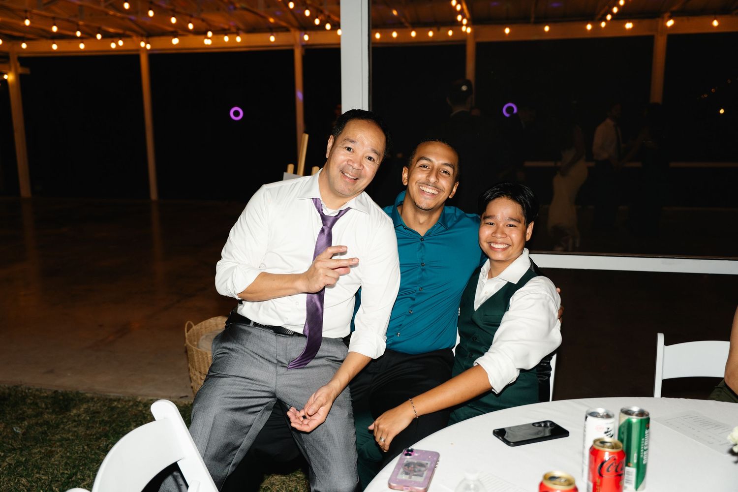 Three smiling wedding guests pose together at a white round table under string lights.