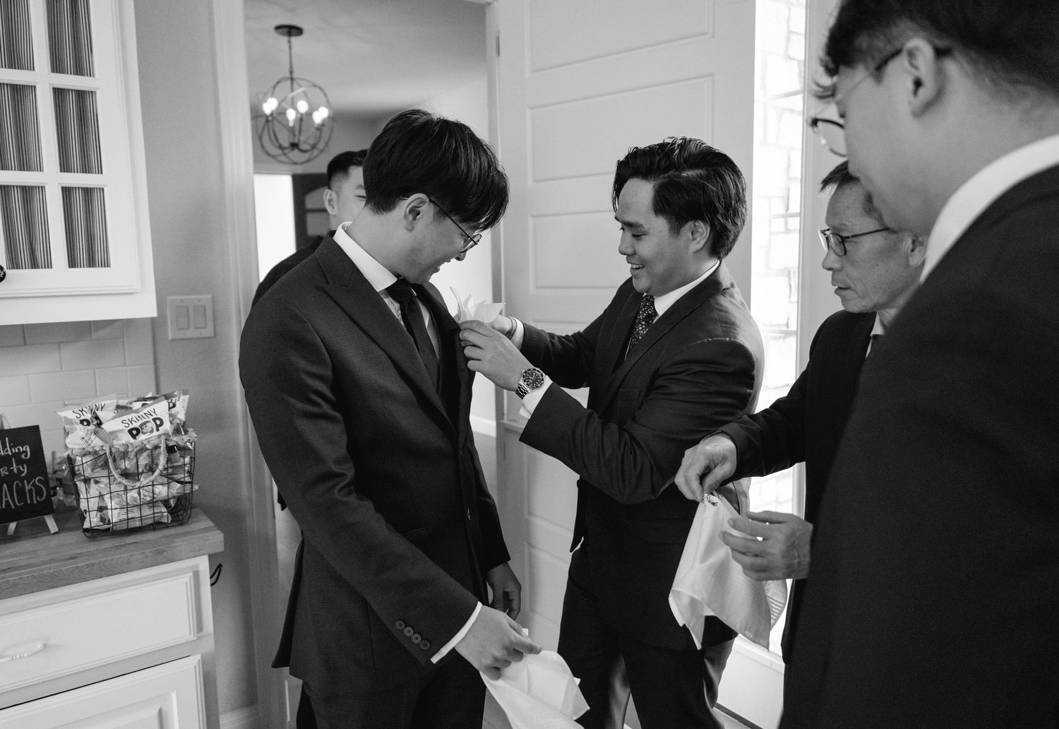 Groomsmen helping groom with boutonniere in black and white wedding preparation photo.