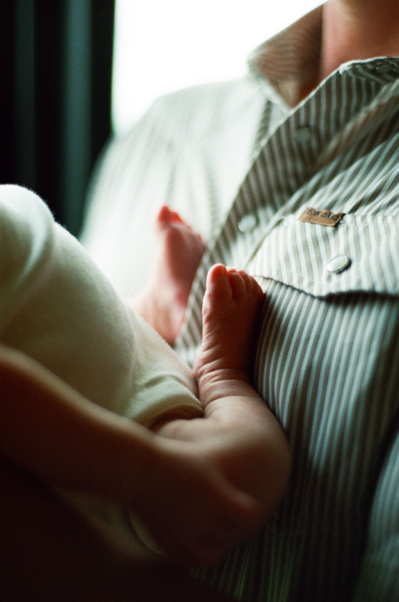 Close-up of a newborn baby's tiny feet resting against an adult's striped button-up shirt.