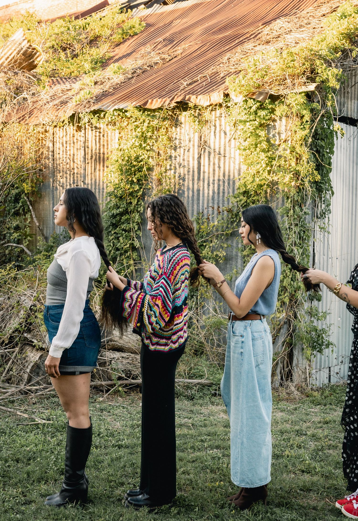Three women standing outdoors near a rustic corrugated metal wall covered in vines, styling each other's hair.