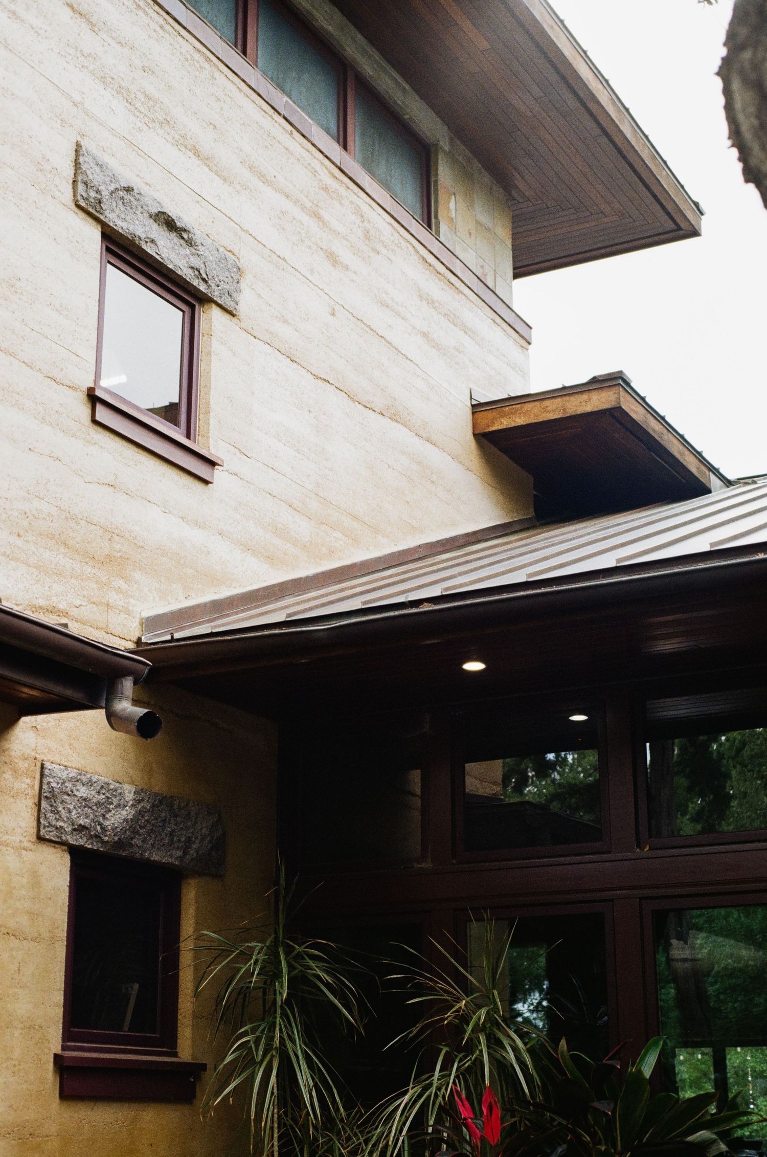 Multi-story Frank Lloyd Wright-style home featuring limestone walls, dark wood trim, metal roof, and tropical plants at entrance.
