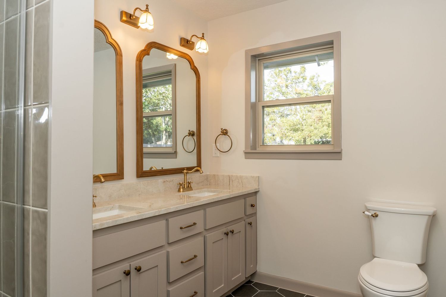 Updated bathroom with gray vanity, marble countertop, arched gold-framed mirrors, and natural window light.