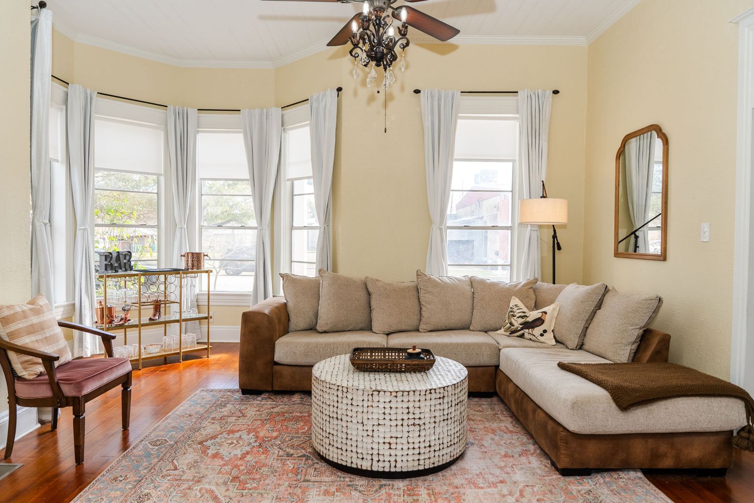 Bright living room with bay windows, sectional sofa, woven ottoman, hardwood floors, and decorative area rug.