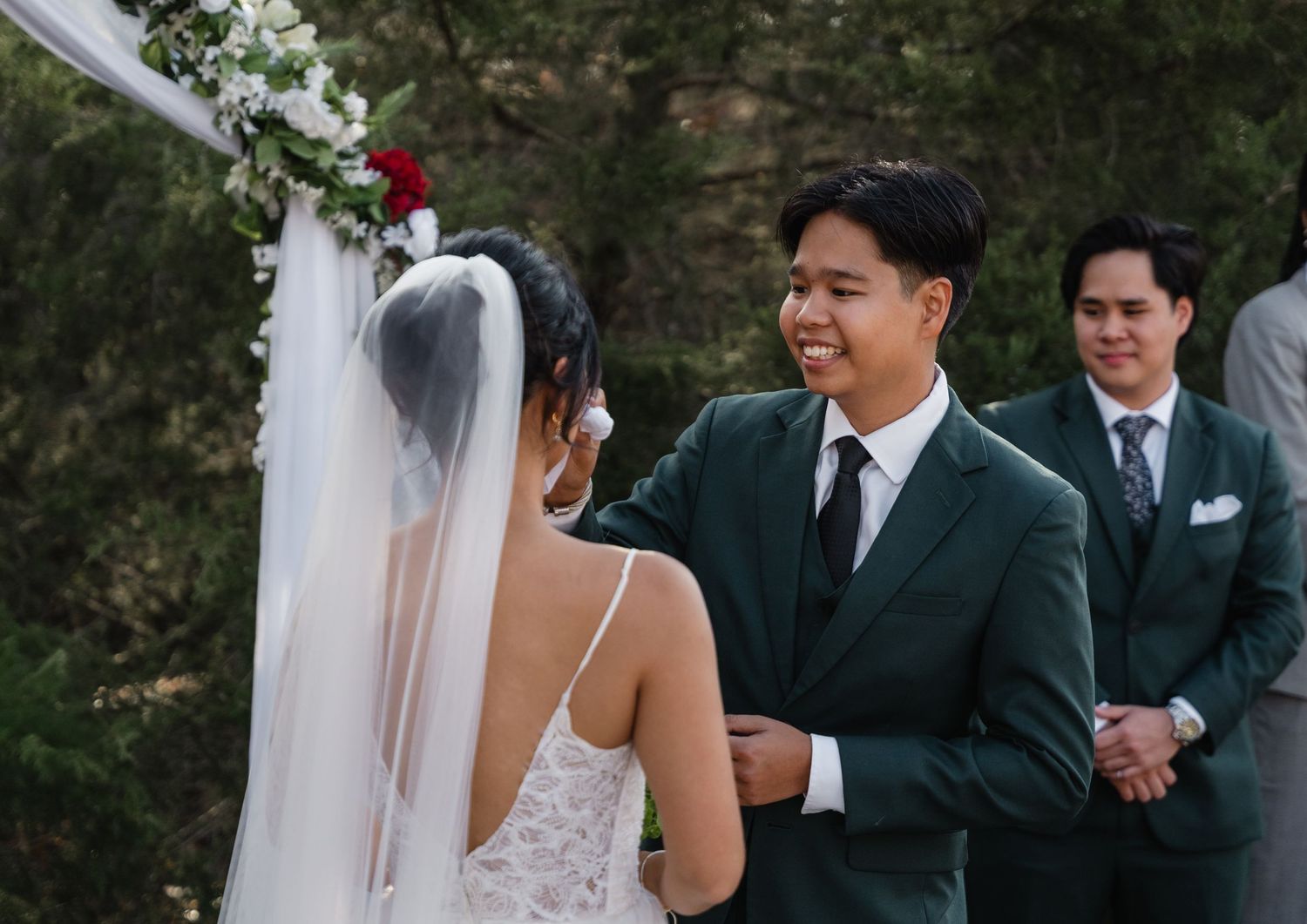 Groom in dark green suit smiles at bride during outdoor wedding ceremony under floral arch.