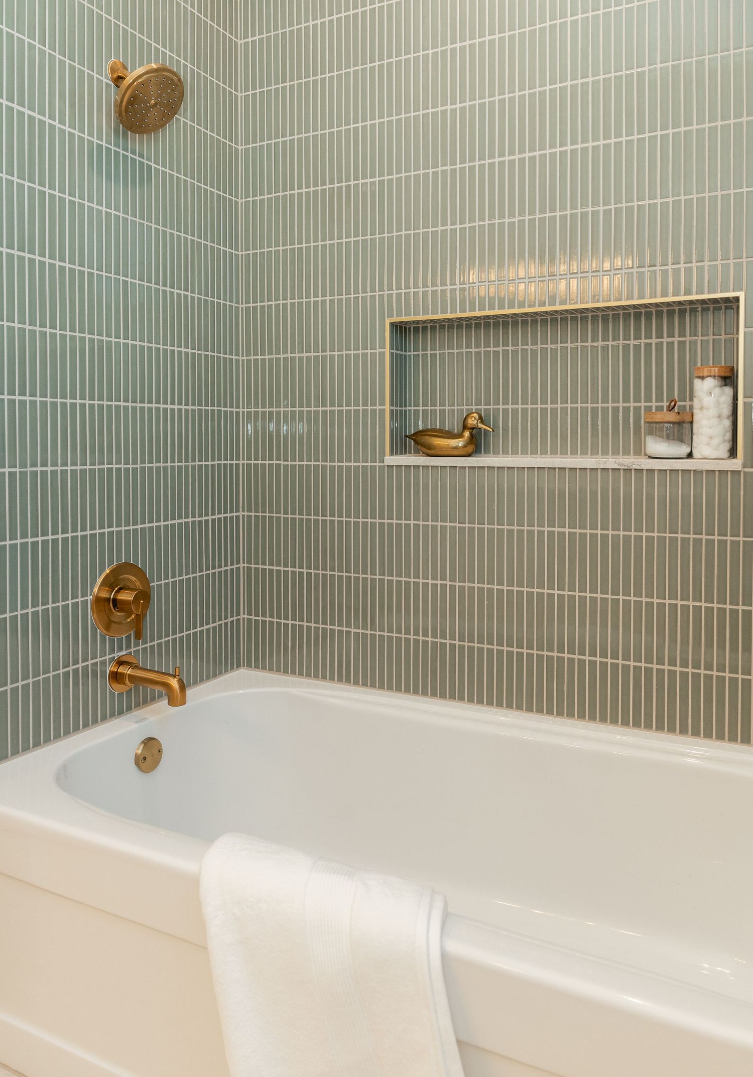 Elegant bathroom with sage green vertical subway tiles, gold fixtures, built-in niche shelf, and white soaking tub with folded towel.