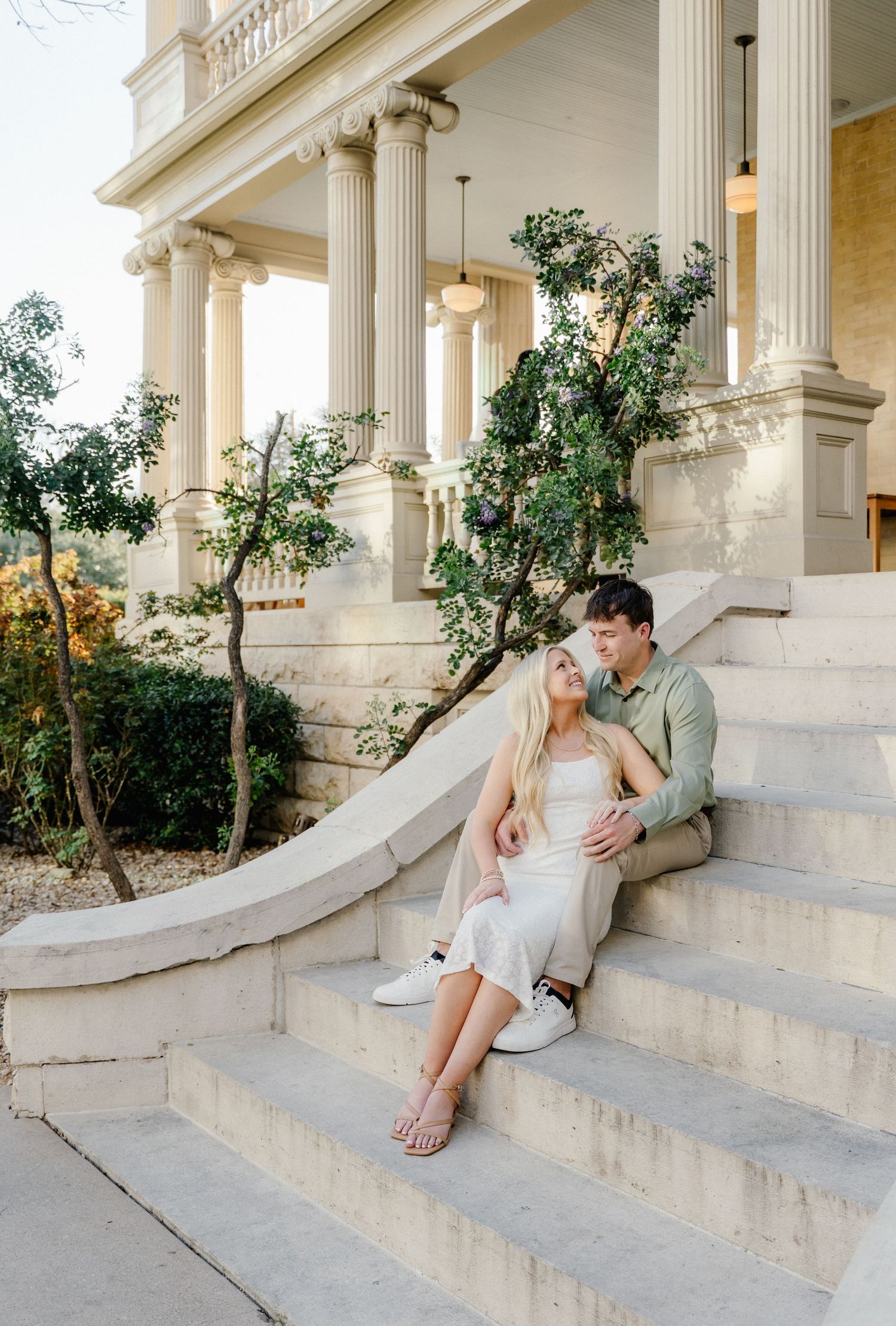 Couple sitting closely on curved stone steps of elegant columned building, sharing a tender moment.