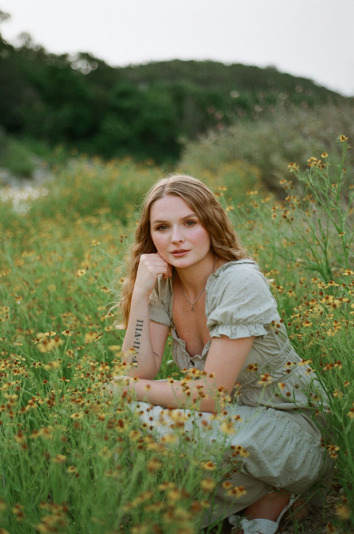 Young woman in sage green dress crouching among wildflowers in a field, posing for a portrait photo.