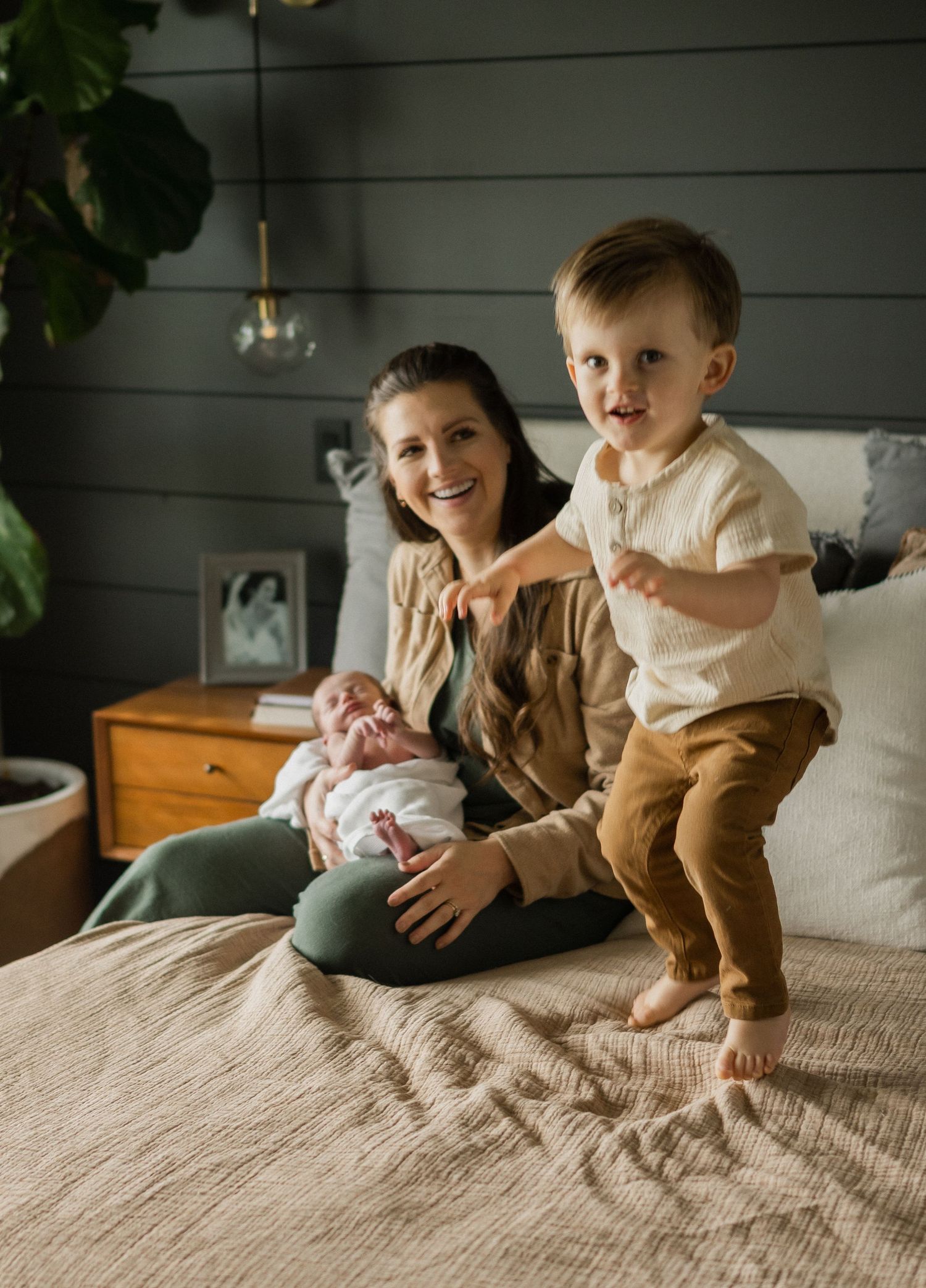 Smiling mother holding newborn baby on bed while energetic toddler boy stands beside her.