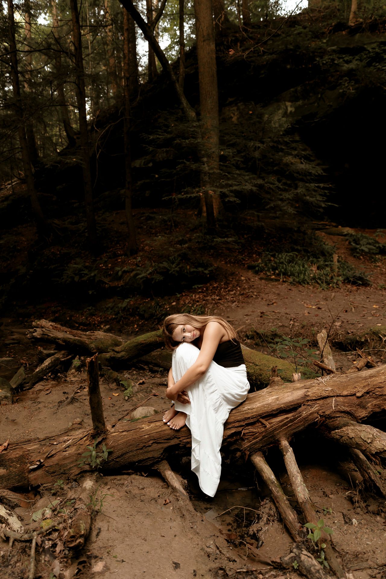 Woman in black top and white skirt poses on fallen logs in a dark forest setting.