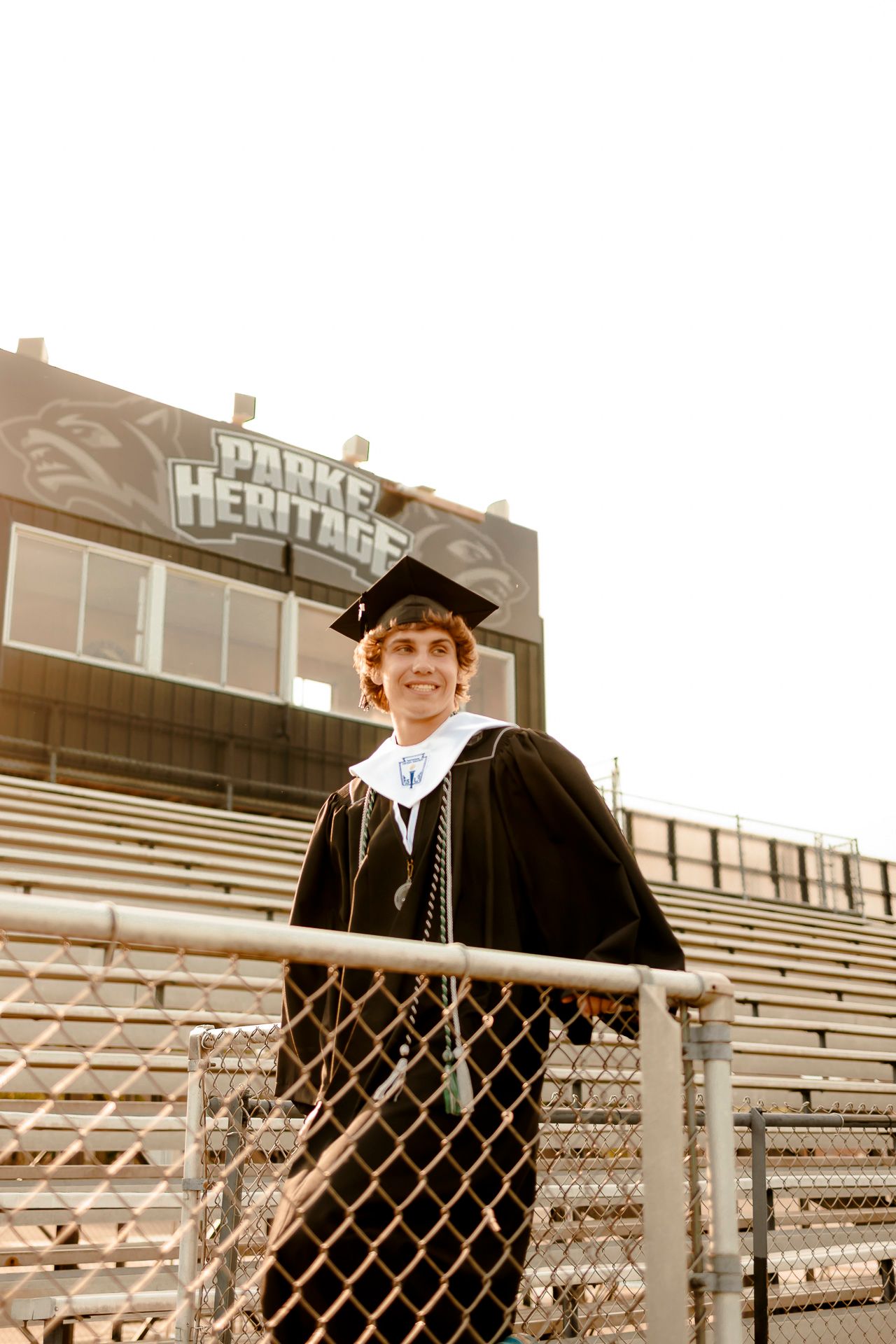 Smiling graduate in black cap and gown stands at Parker Heritage stadium bleachers, posing by chain-link fence.