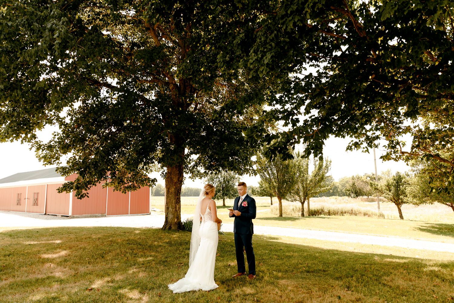 Bride and groom share private vows under a large oak tree on a sunny day with a red barn in the background.