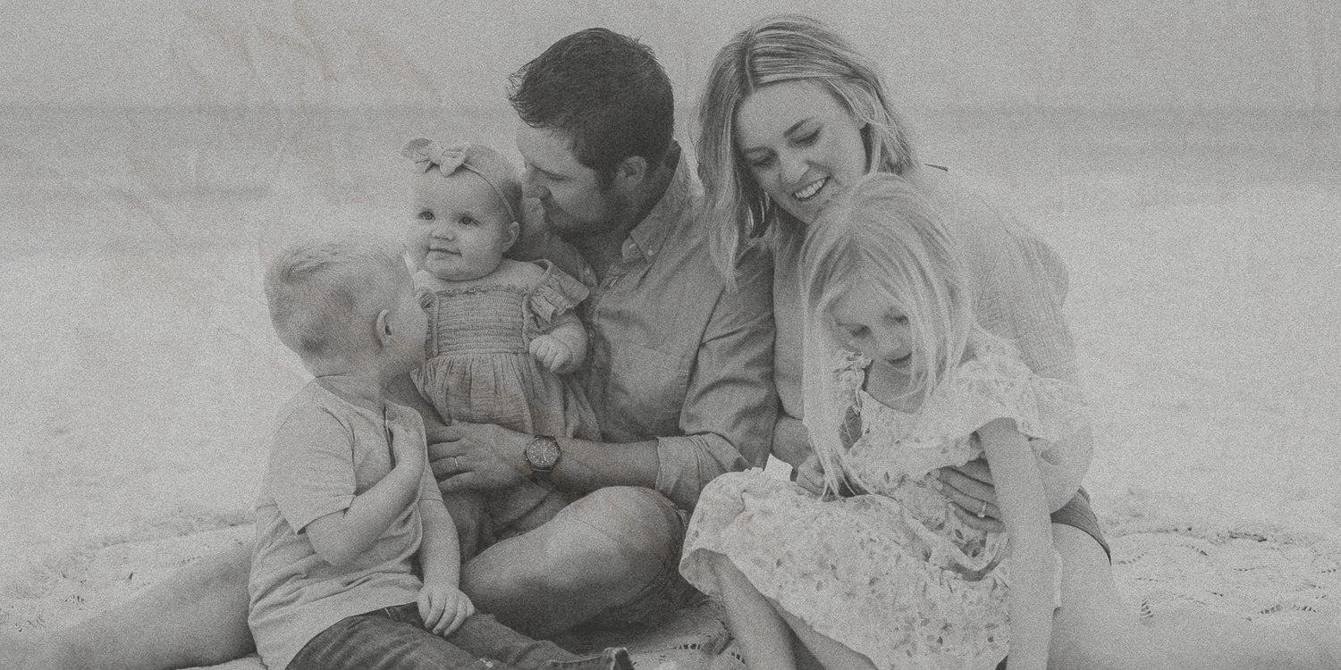 A family sits together on a beach in black and white, creating an intimate moment in soft natural lighting.