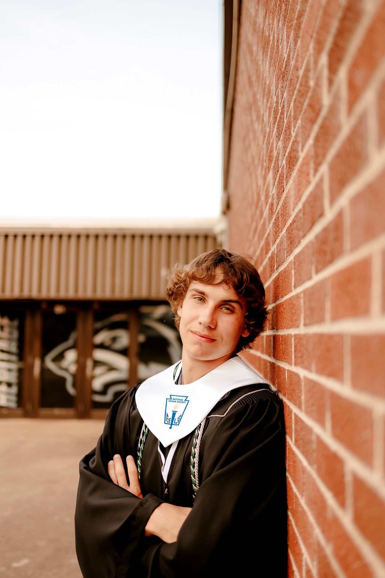 Young man in graduation gown and stole leans against red brick wall with arms crossed, smiling confidently.