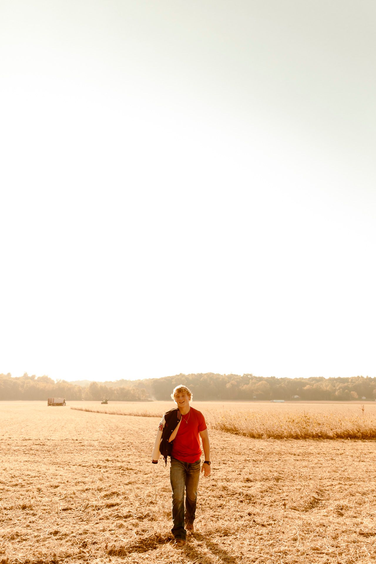 Man in red shirt walking through golden field at sunset, carrying camera bag in warm backlit countryside.