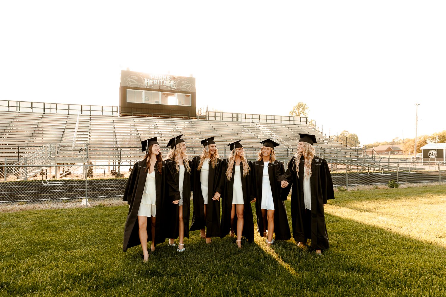 Group of female high school graduates in black caps and gowns posing on football field at golden hour.