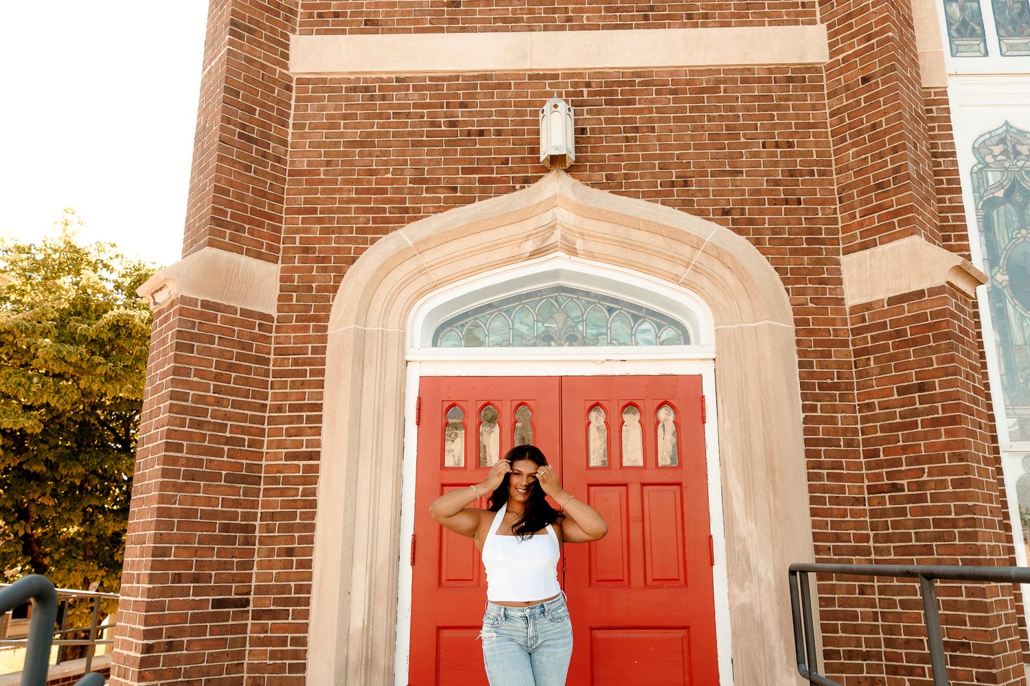 Woman in white top and jeans posing before a red arched door on a brick building exterior.