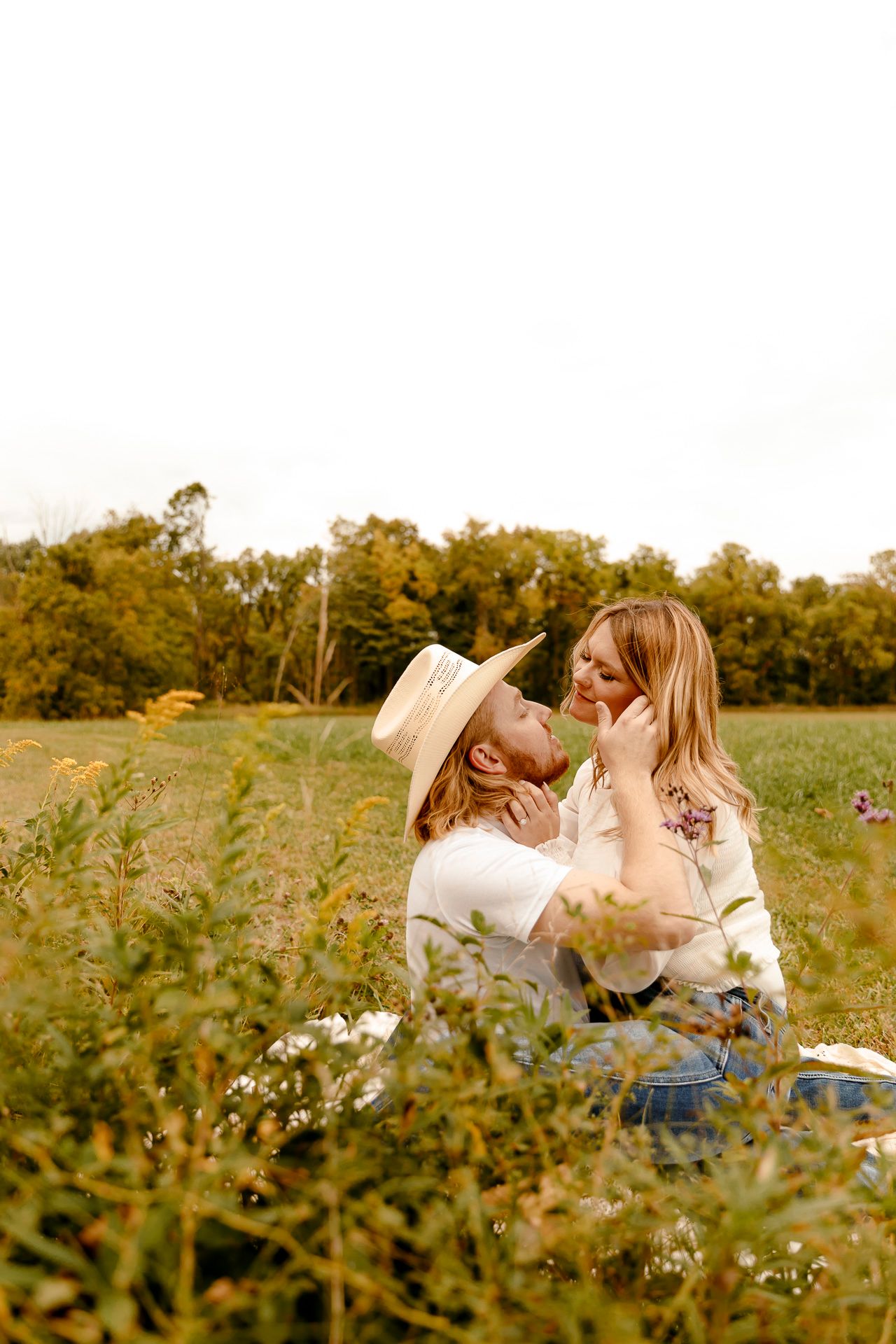 Couple sharing a romantic moment in a sunlit meadow, man in straw hat and woman in floral top sitting in green grass.
