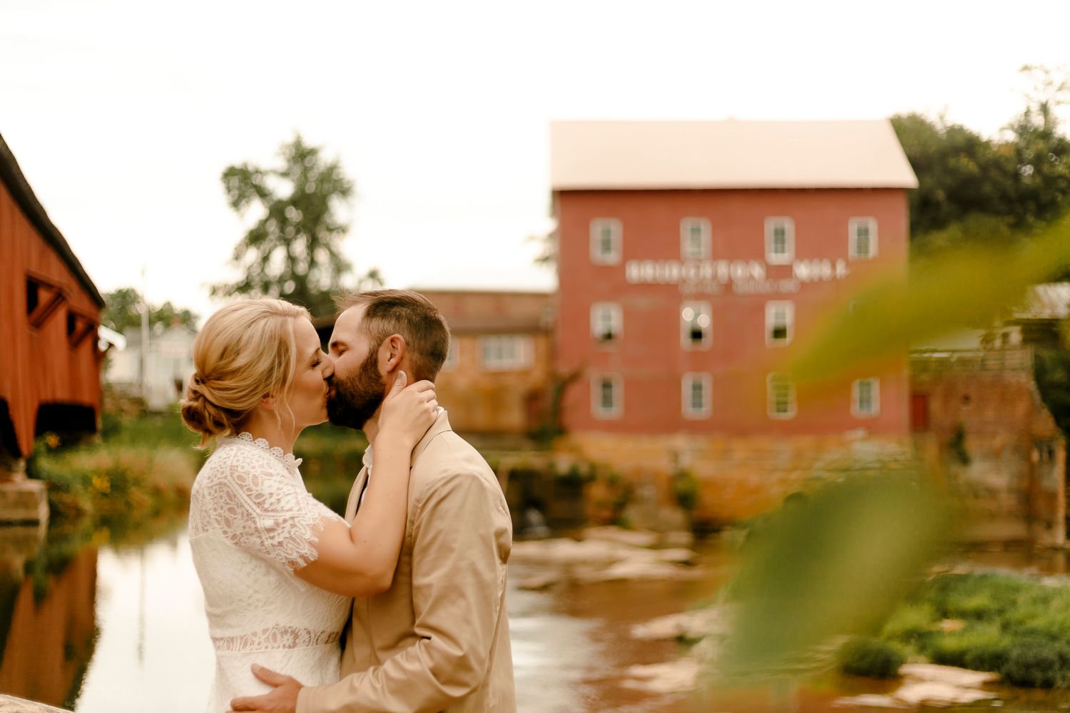 Bride and groom sharing a kiss near a covered bridge with Brighton Mill in the background.
