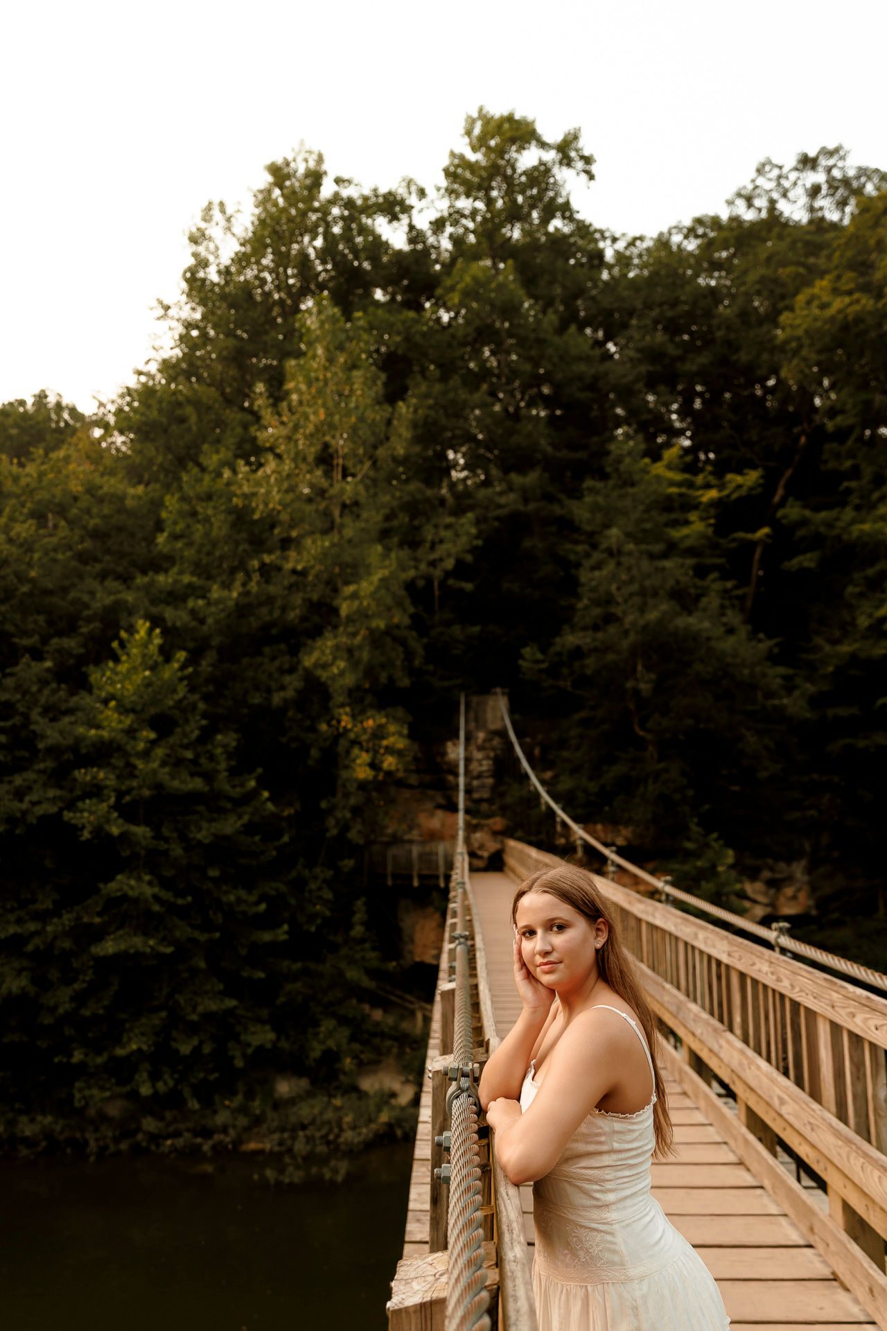 Young woman in white dress posing on a wooden suspension bridge surrounded by lush green trees over a river.