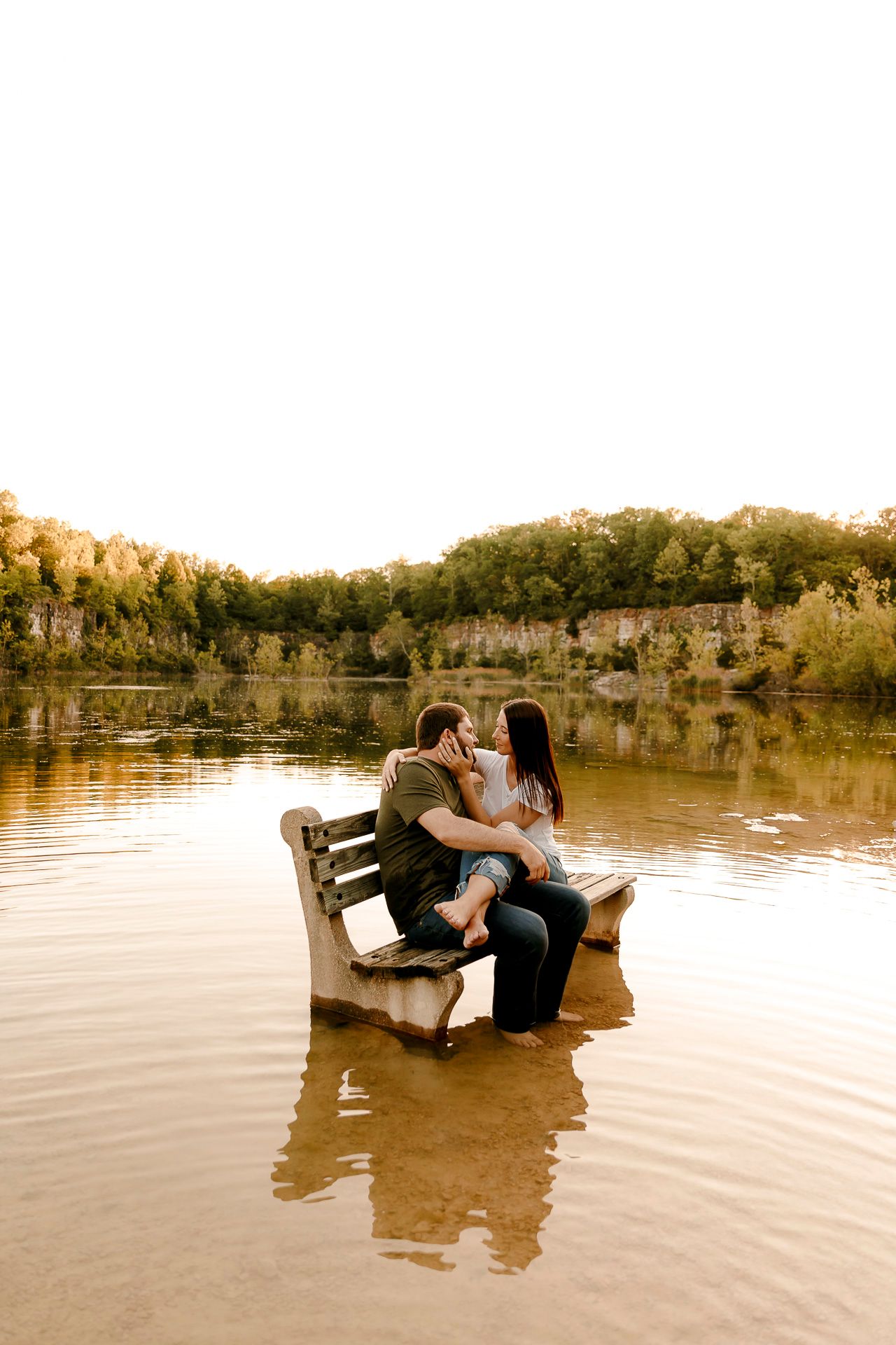 Couple sitting closely on a wooden bench submerged in a calm lake, surrounded by lush trees at golden hour.