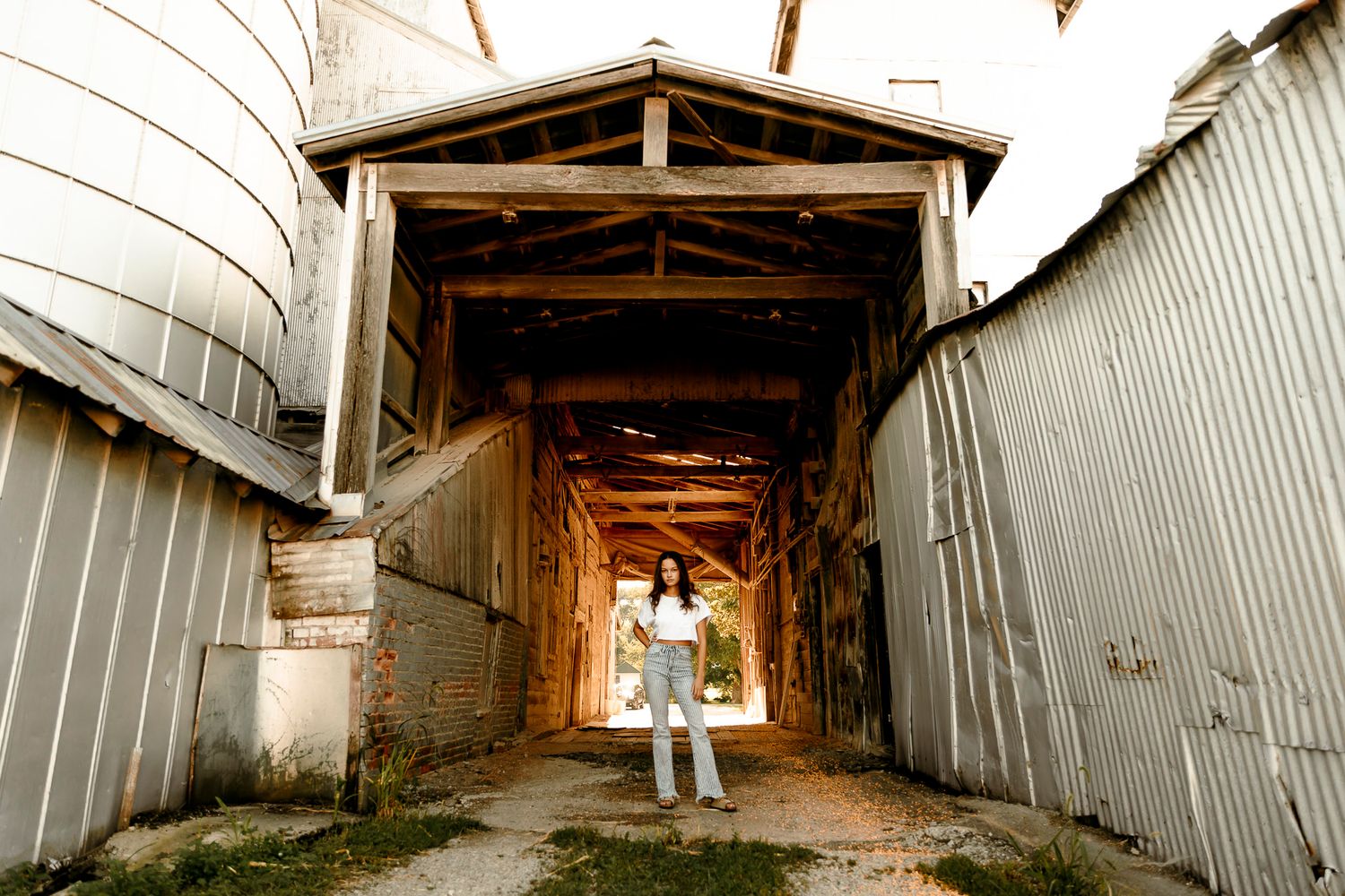 Young woman in jeans stands in rustic barn corridor flanked by corrugated metal silos and wooden beams.
